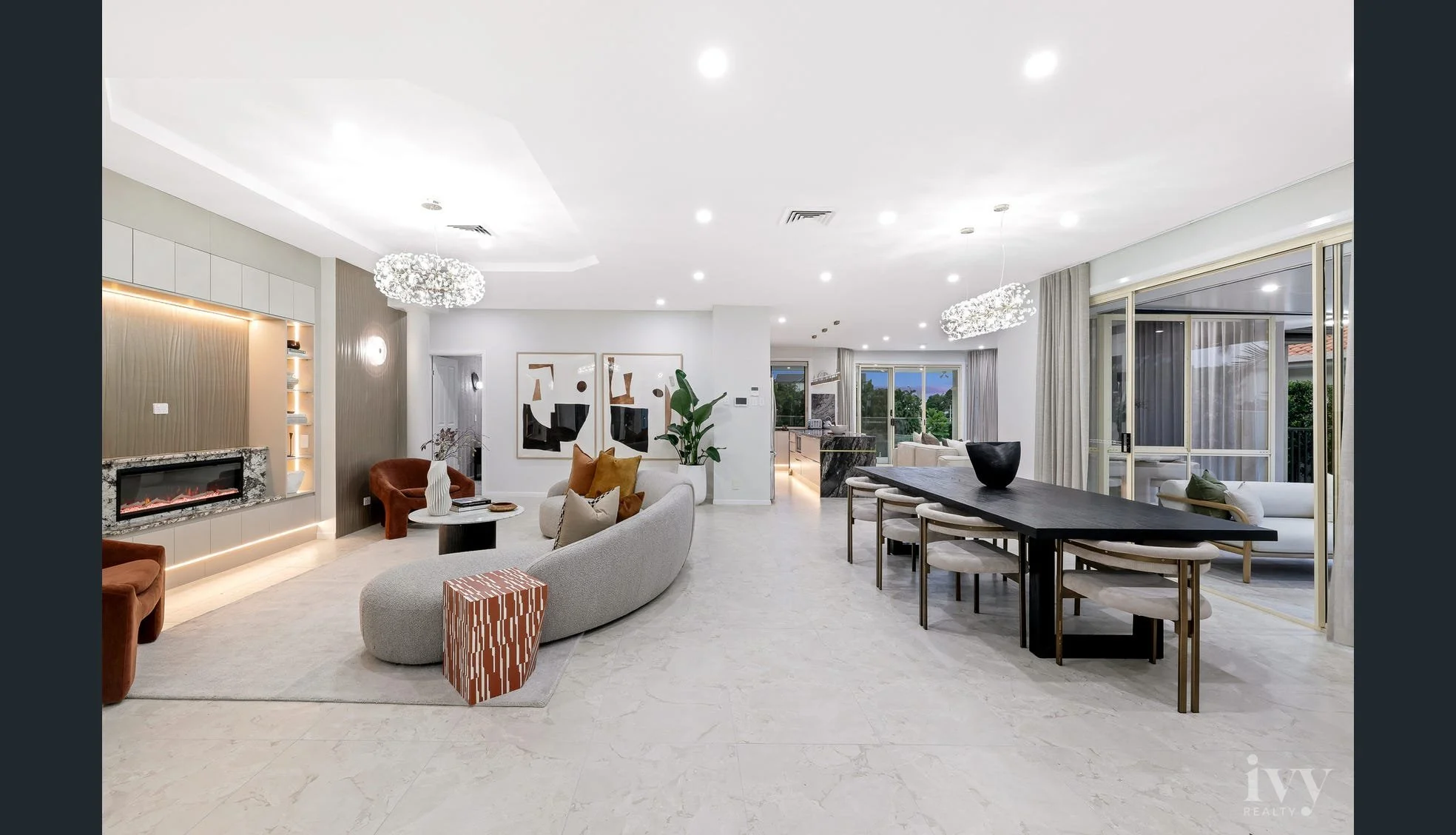 Spacious modern living room with white walls, light-colored flooring, a fireplace, and various seating options including a sofa and armchairs. Elegant lighting fixtures and large windows with curtains offer natural lighting and outdoor views.