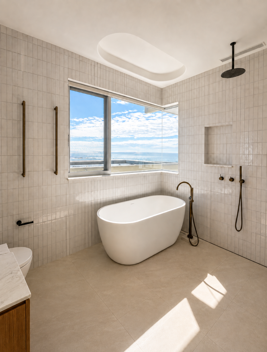 Modern bathroom with a freestanding bathtub, large window showing ocean view, beige tiled walls and floor, rain shower, and gold bathroom fixtures.