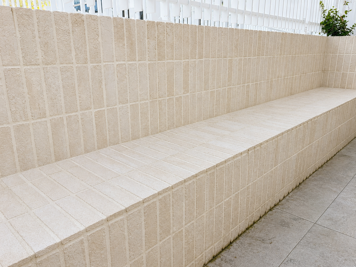 Long beige tiled bench with textured surface against beige tiled wall, outside area with white fence and green plant in background.