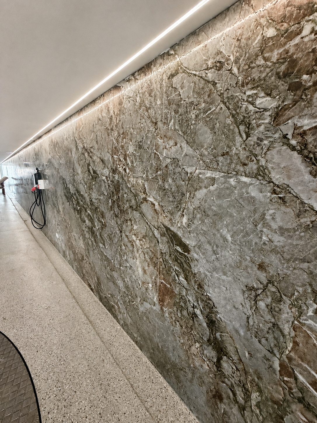 Close-up view of a large, polished marble wall with intricate veining in shades of gray, brown, and white. The wall has a subtle strip of lighting along the top edge, and part of the floor is visible, showing a textured surface. There is some equipme
