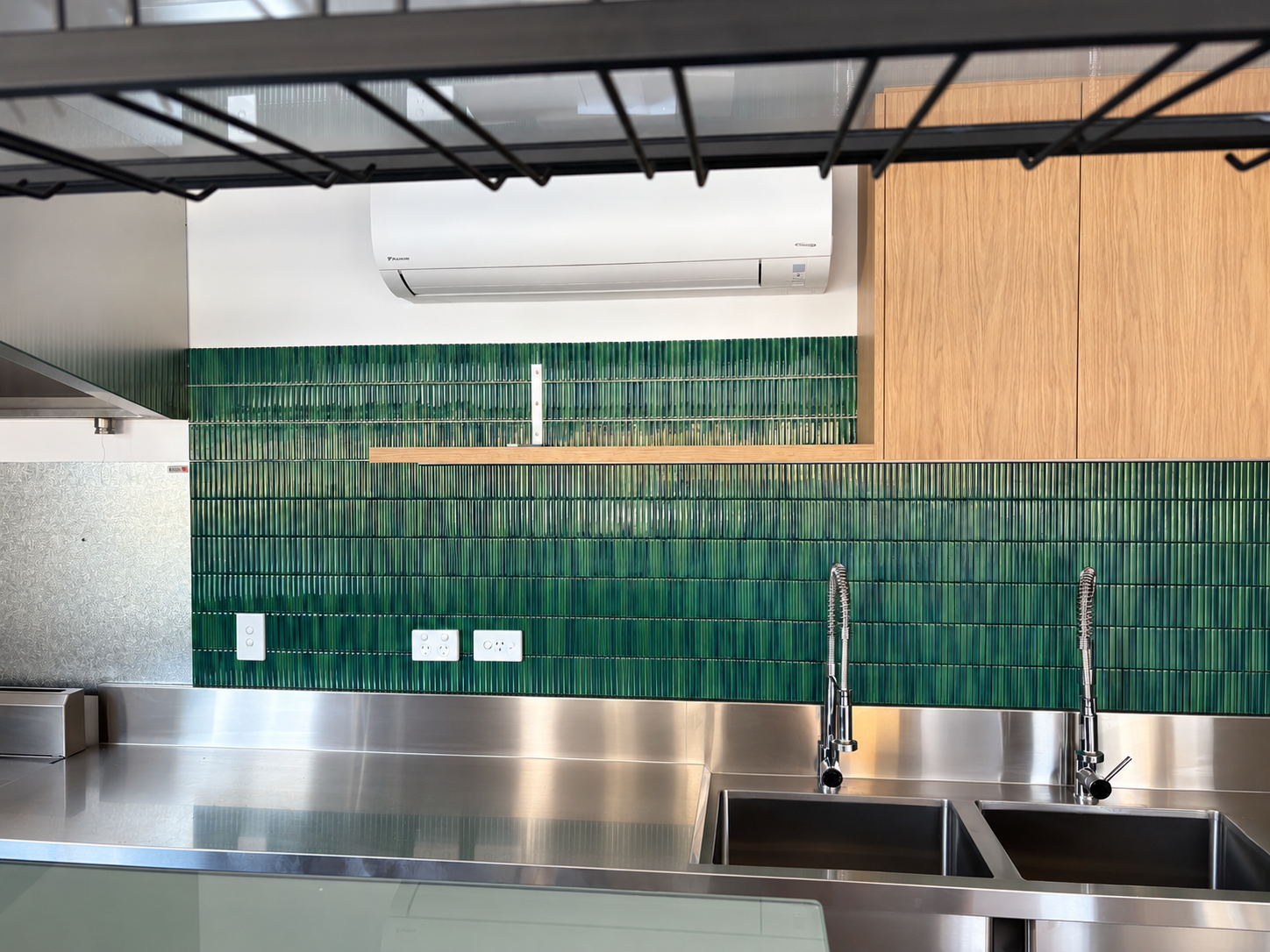 Kitchen with stainless steel sink, green tiled backsplash, wooden cabinets, wall-mounted air conditioner, and electrical outlets.