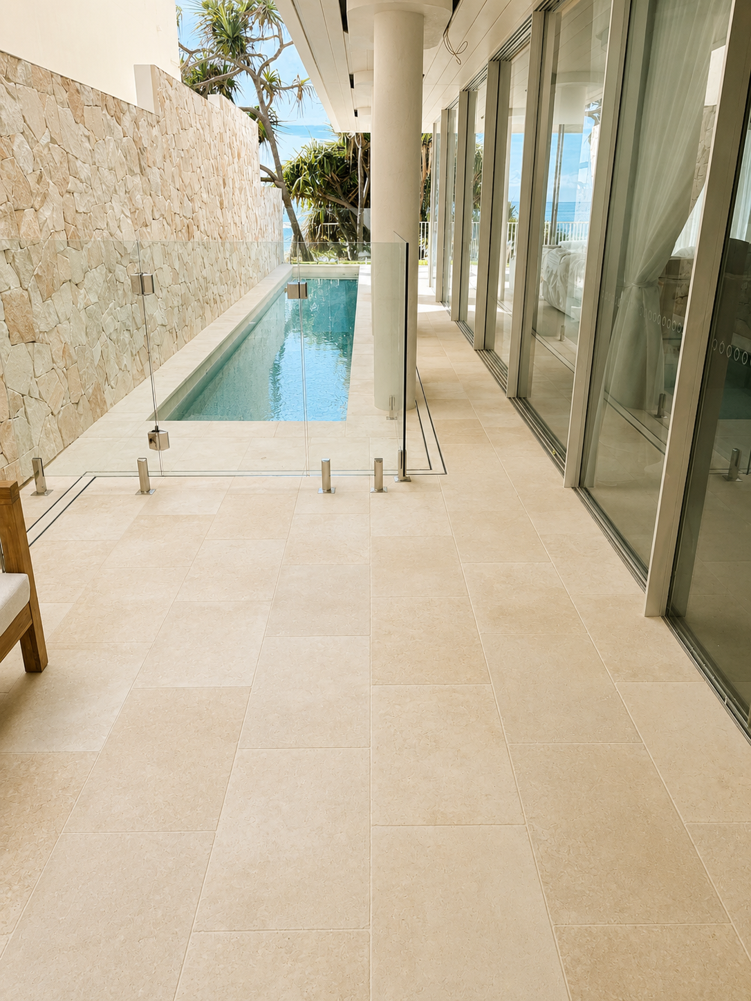 Modern outdoor patio with a small rectangular swimming pool, glass fence, beige tiled floor, stone wall, and sliding glass doors leading inside, with palm trees and blue sky in the background.