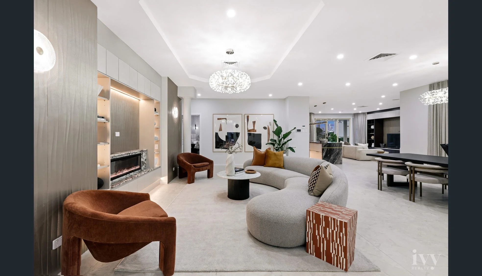 Modern living room with beige curved sofa, orange and patterned throw pillows, brown velvet armchair, white vase with plants on coffee table, contemporary art on wall, white ceiling with chandelier, and open layout connecting to dining and kitchen ar