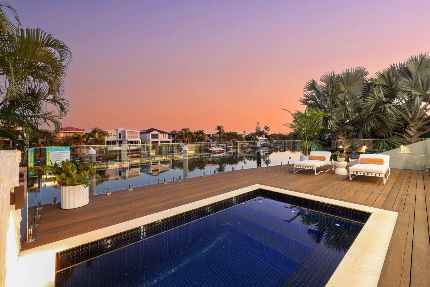 A luxurious rooftop deck with a small swimming pool, two white lounge chairs with orange cushions, potted plants, and a glass railing overlooking a canal with modern houses at sunset.