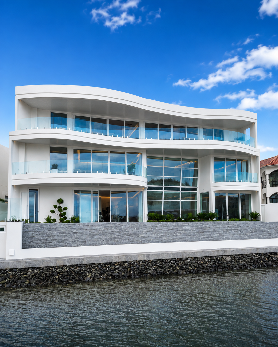 Modern white multi-story waterfront house with glass balconies and large glass windows, surrounded by landscaping with small trees and plants, under a partly cloudy sky.