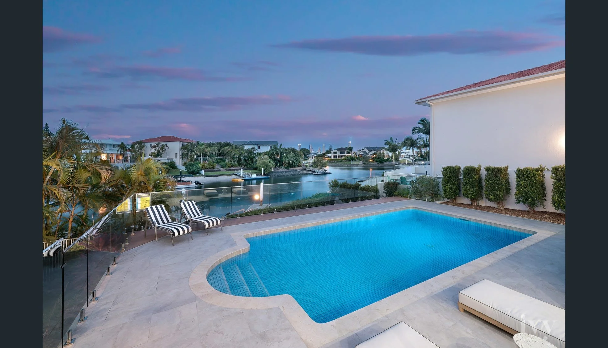 Swimming pool with striped lounge chairs overlooking a canal and residential houses at dusk.