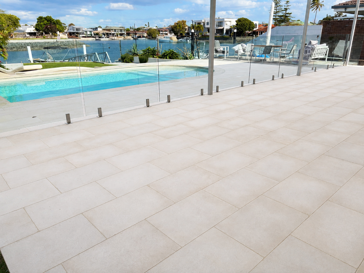 Empty outdoor patio with beige tiles, glass safety fence, swimming pool, lounging chairs, and a waterway with boats and houses in the background under a partly cloudy sky.