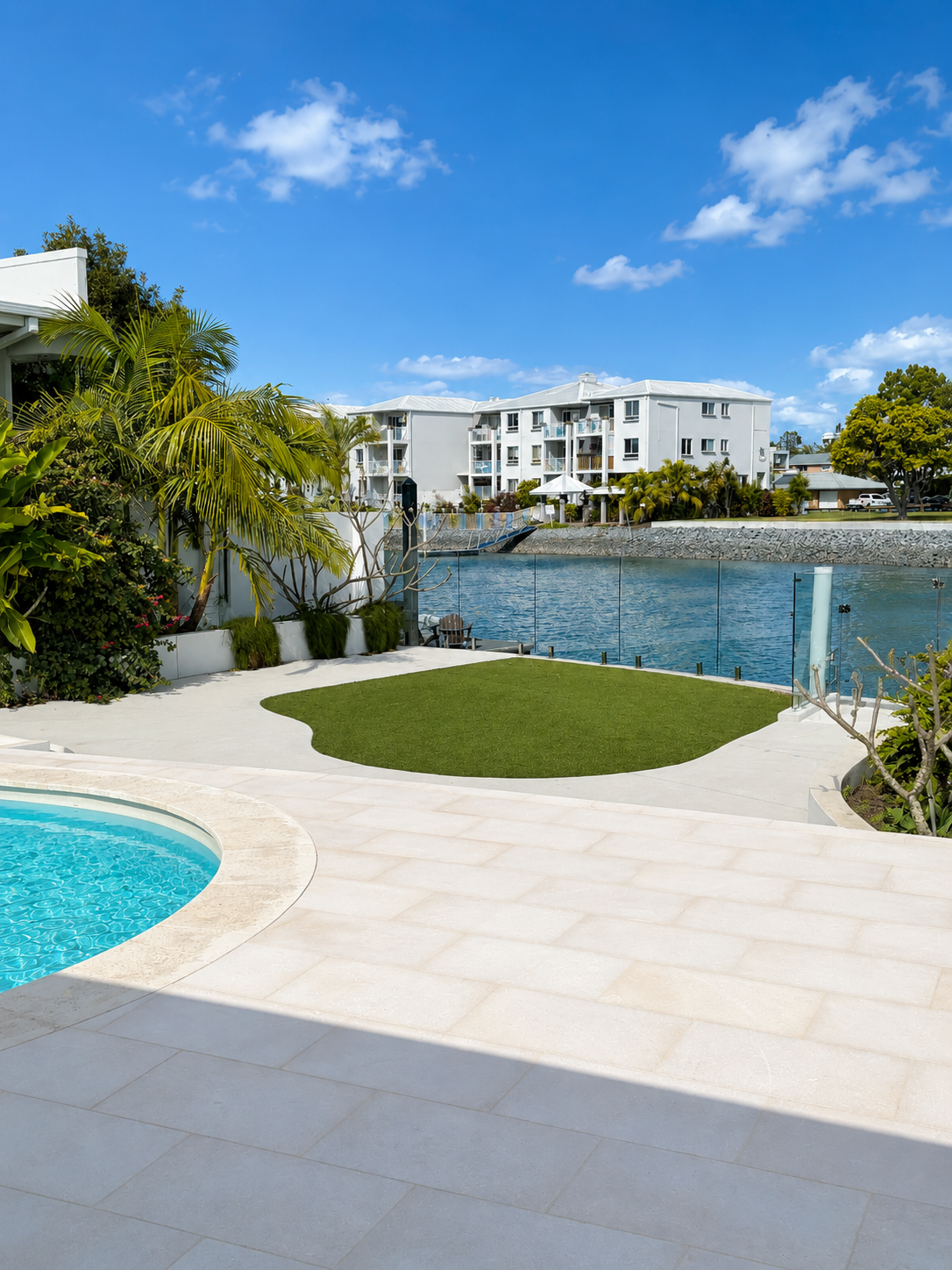 View of a modern poolside area with a small pool, tropical plants, and a grassy patch, overlooking a body of water with residential buildings across under a clear blue sky.