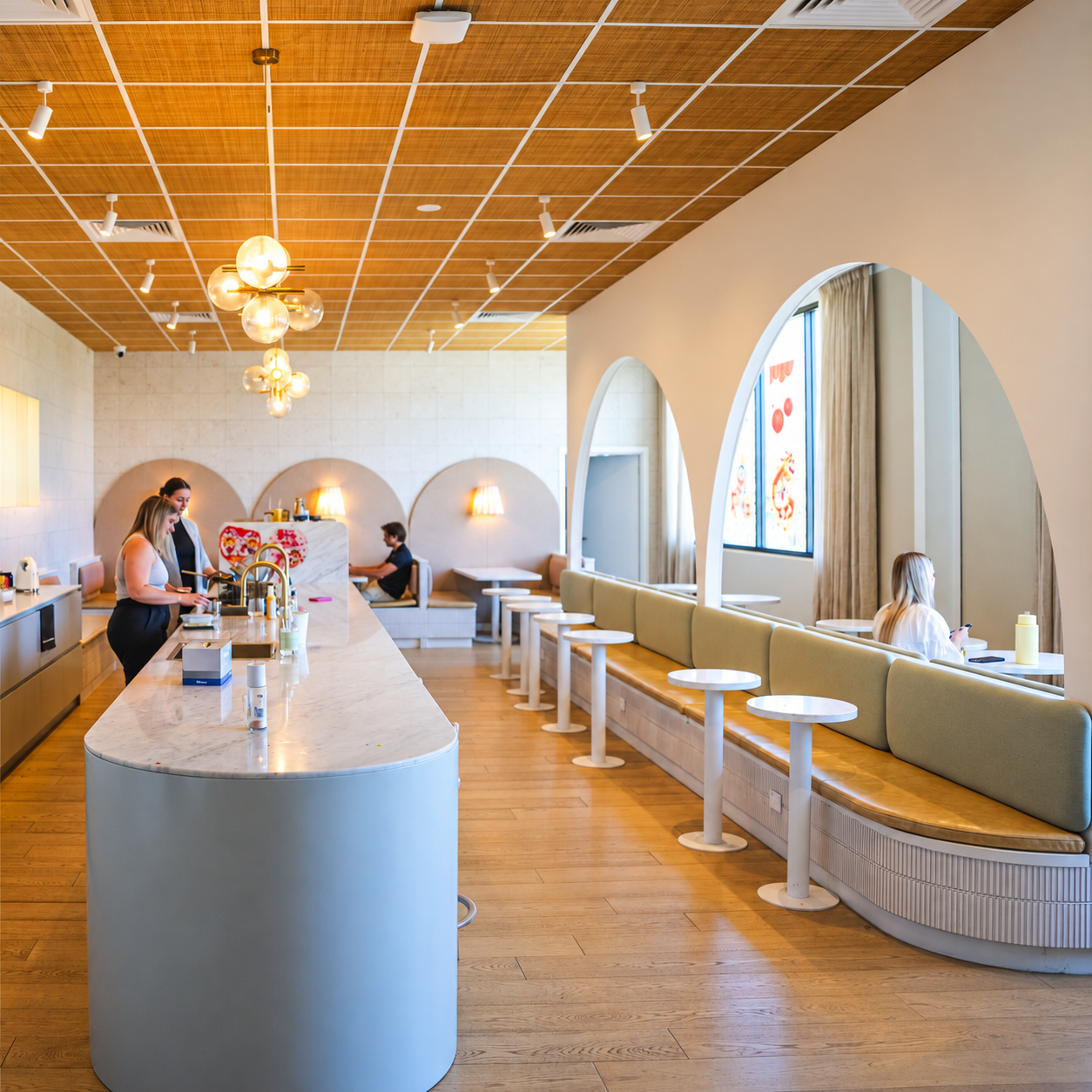 Interior of a modern cafe with a white marble counter, wooden floor, beige seating with small round tables, large windows with curtains, and decorative lighting fixtures.