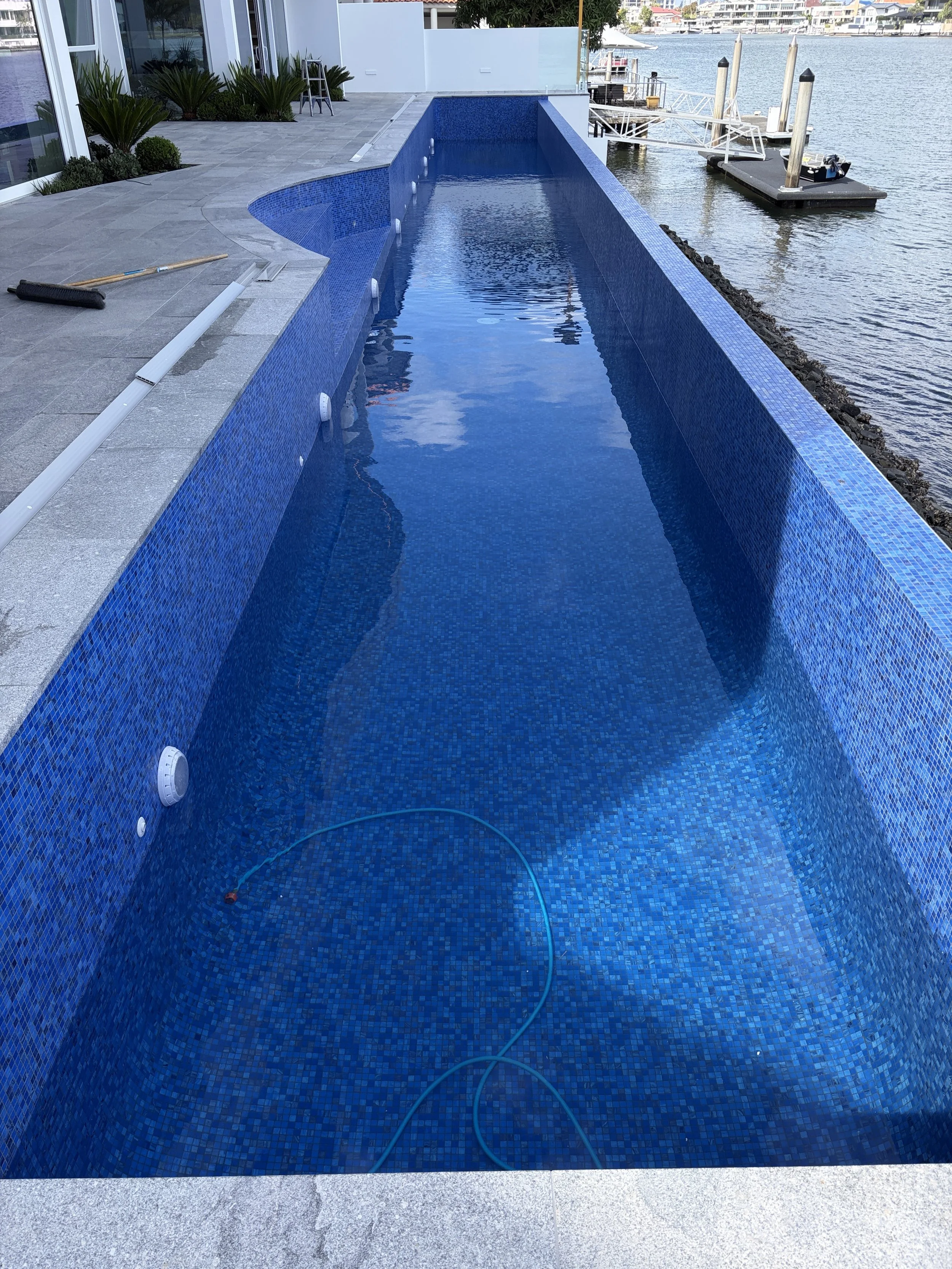 An empty rectangular swimming pool with blue mosaic tiles, located on a patio next to a body of water, with a dock and boats visible in the background.