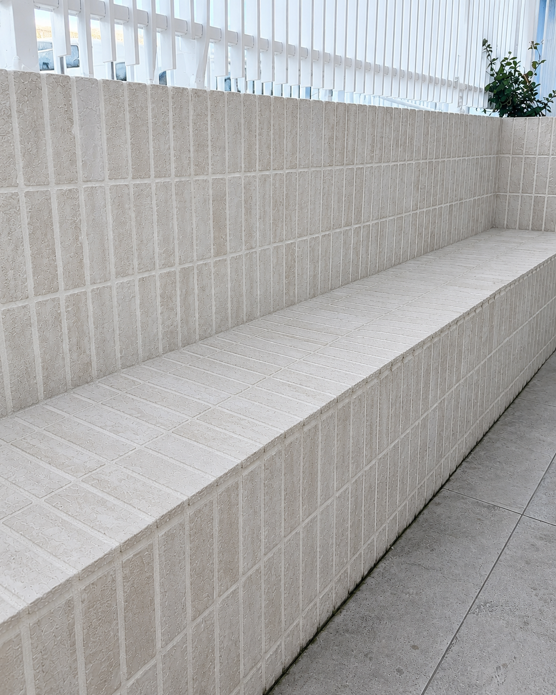 A white tiled bench with a textured surface, located indoors, with a plant and windows in the background.