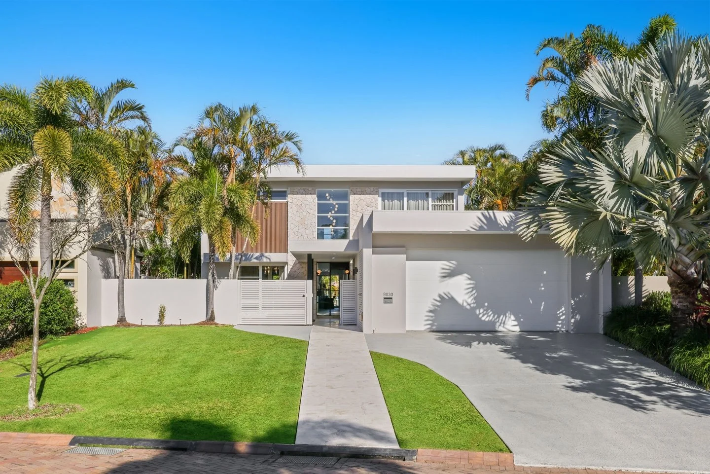 Modern two-story house with white exterior, large windows, surrounded by palm trees and greenery, with a concrete driveway and walkway, under a clear blue sky.