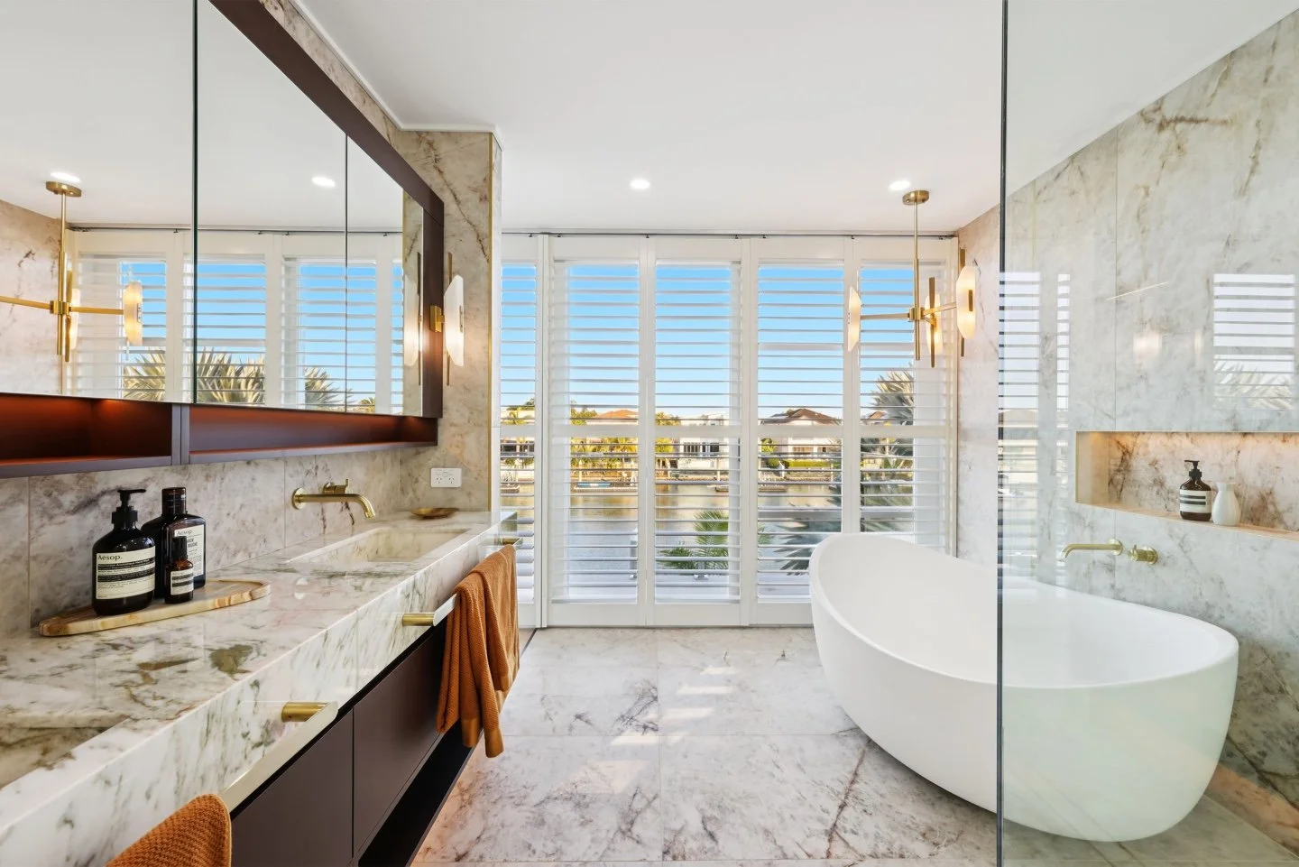Modern bathroom featuring a freestanding white bathtub, a marble countertop with a sink, large windows with white plantation shutters, and marble walls and floors.