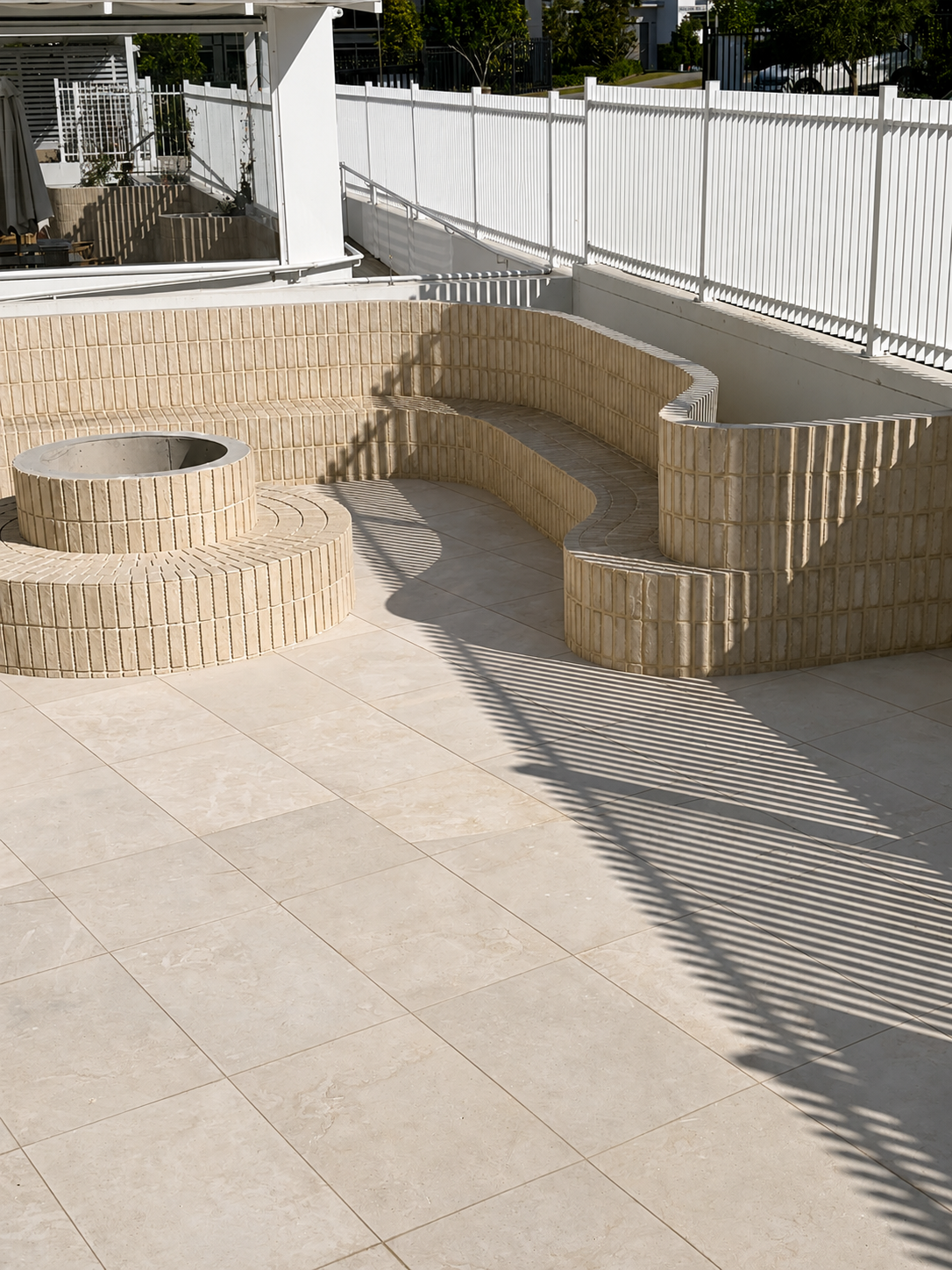 Outdoor area with beige tiled seating arranged in circular and curved shapes, surrounded by a white fence, with shadow patterns from a nearby palm frond cast across the ground.