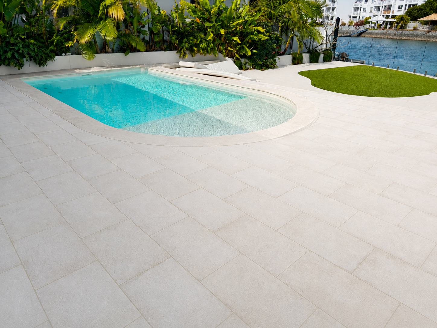 A modern outdoor swimming pool with clear water, surrounded by a white tiled deck. There are lush green plants along the back wall, and a small patch of green grass near the water. In the background, there is a body of water and several white buildin