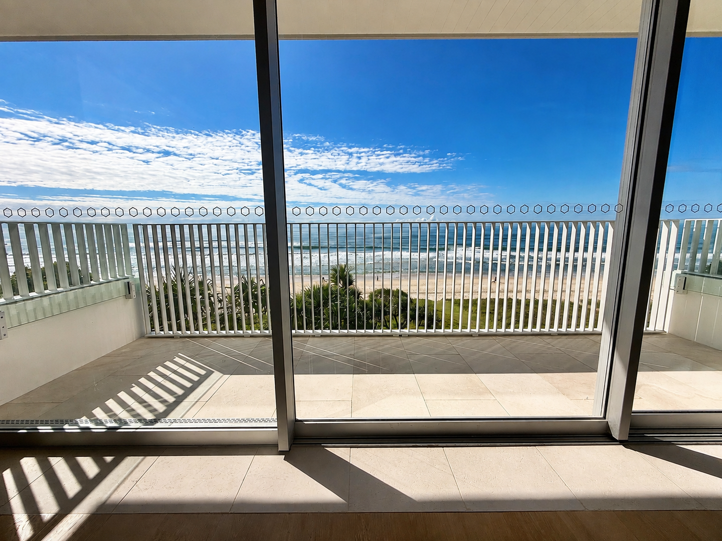 View from a balcony with glass sliding door, overlooking a beach, ocean waves, and sandy shoreline under a blue sky with scattered clouds.
