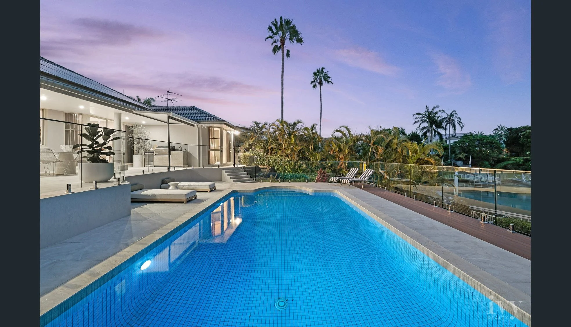 Modern backyard with a swimming pool, lounge chairs, patio furniture, and tropical palm trees at sunset.