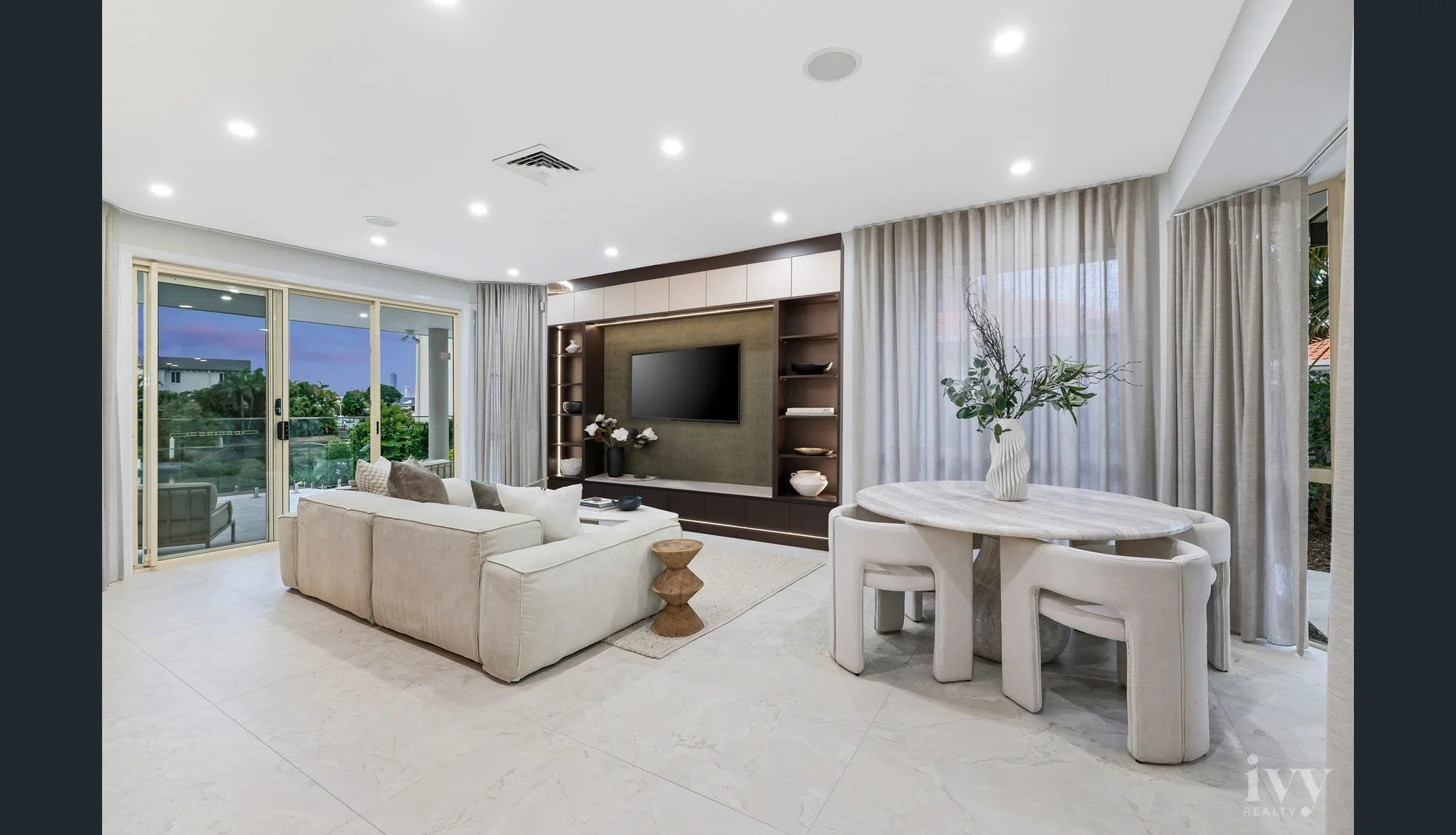 Modern living room with cream sofa, round dining table with white chairs, TV on a stylish wall unit, large windows with sheer curtains, sliding glass door leading to balcony, indoor plants, and minimal decor.