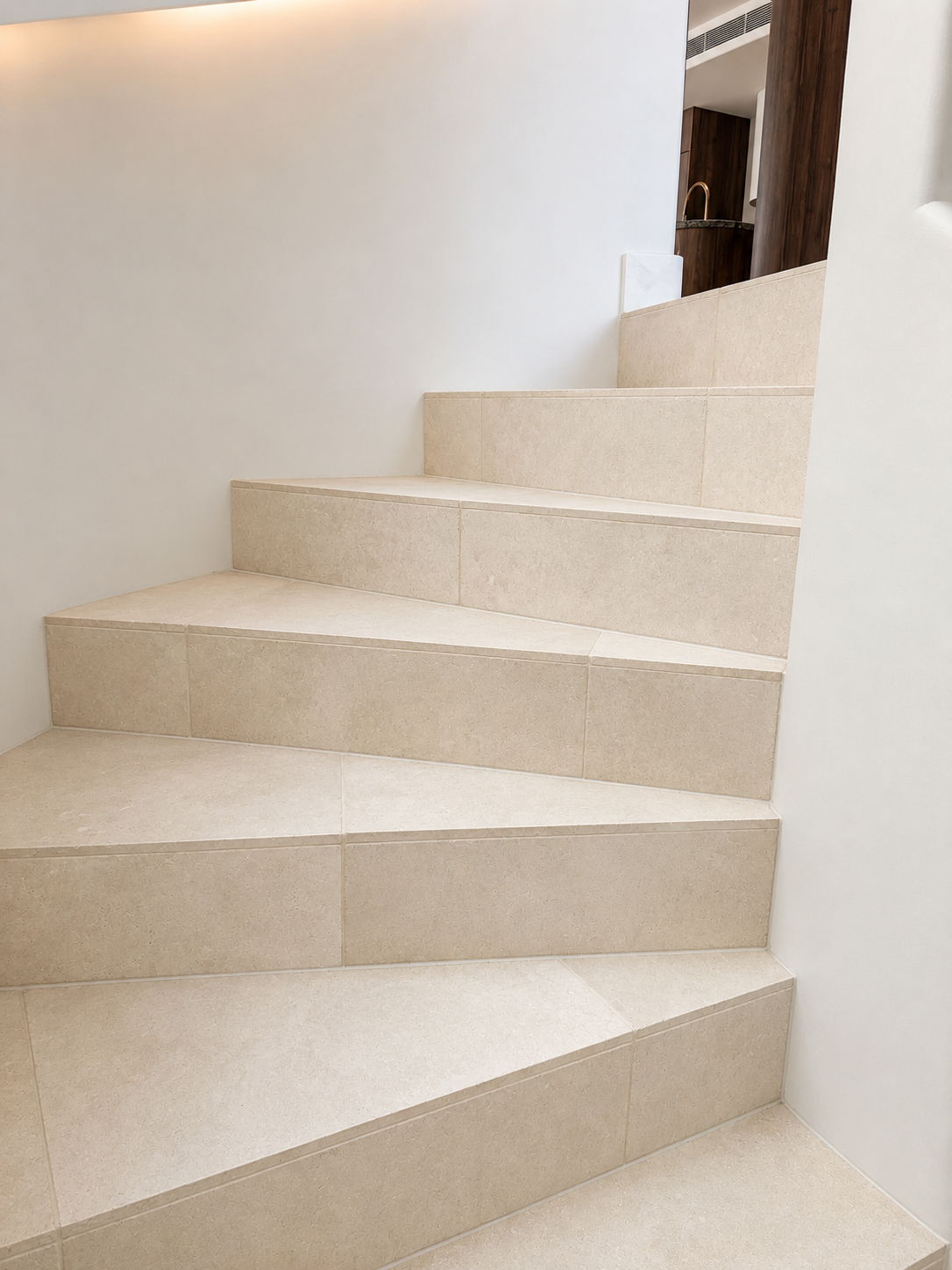 Interior staircase with beige tiles and white walls, leading to a upper floor, with a view of a kitchen area with dark wood cabinets and a gold faucet in the background.