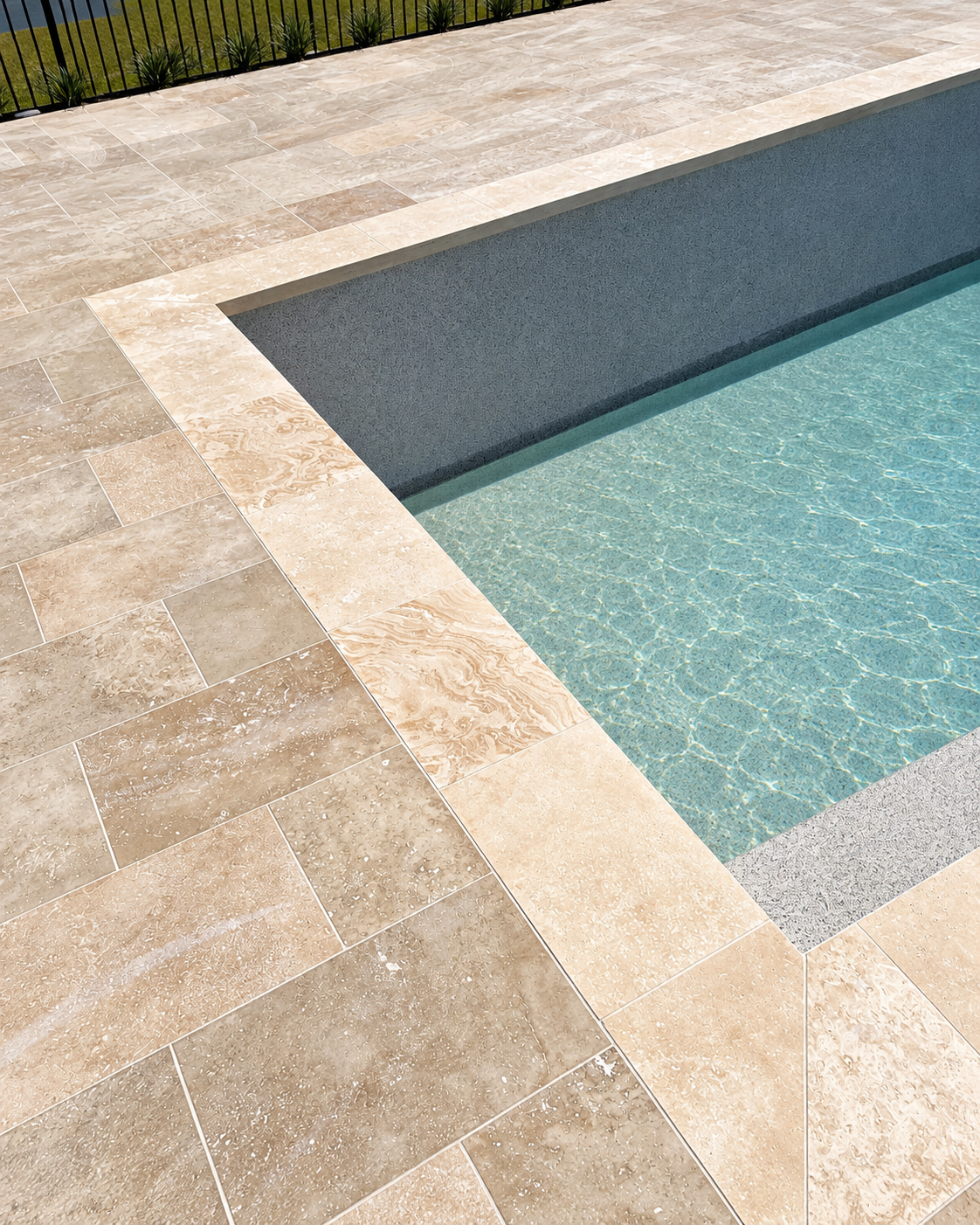 Close-up of a swimming pool with a beige tiled deck and a black metal fence in the background.