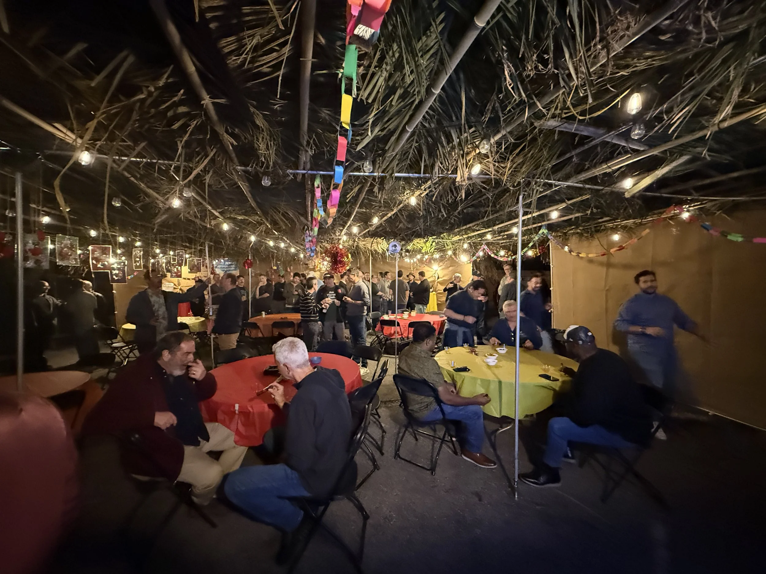 A crowded indoor party or gathering with people sitting and standing around tables covered with colorful tablecloths under a thatched roof decorated with string lights and paper chains.