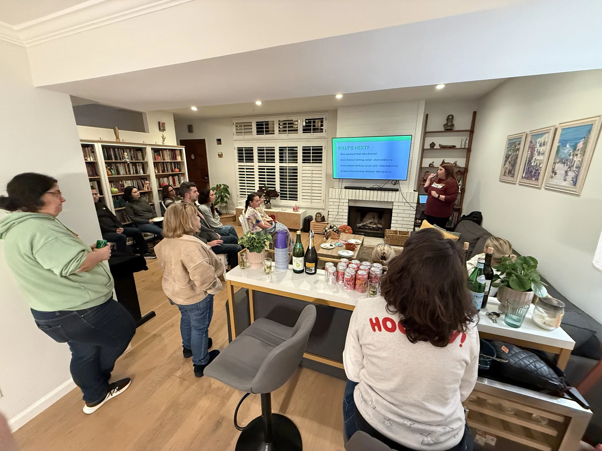 People attend a presentation or social gathering in a cozy living room with a large television screen on the wall, bookshelves, and artwork. Some people are sitting, some are standing, and a woman is speaking near the fireplace.