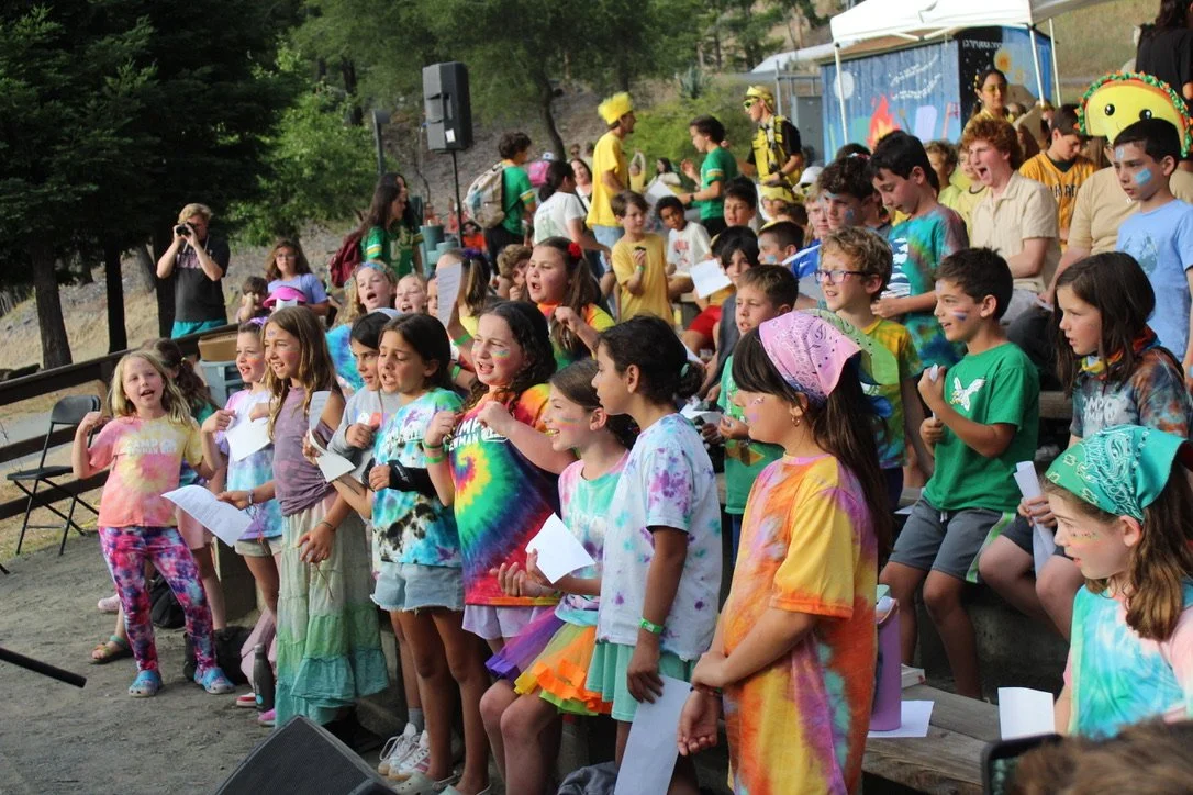 A group of children and teenagers dressed in colorful tie-dye shirts, gathered outdoors, singing or performing together on a stage during a summer event or festival. Some are holding papers, and there are adults and a person in a taco hat in the background.