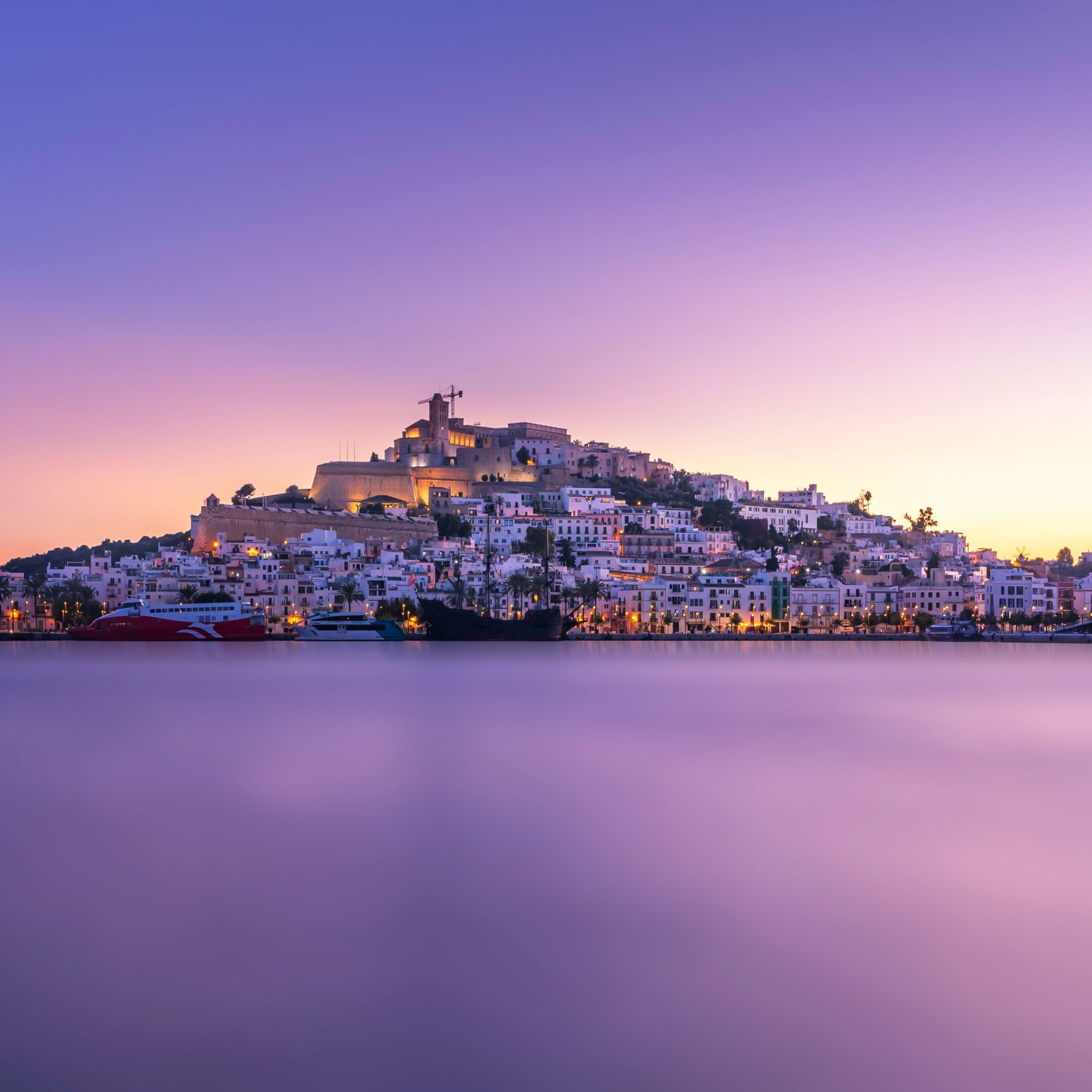 A hillside town with white buildings and a castle at the top, reflected on calm water during twilight.