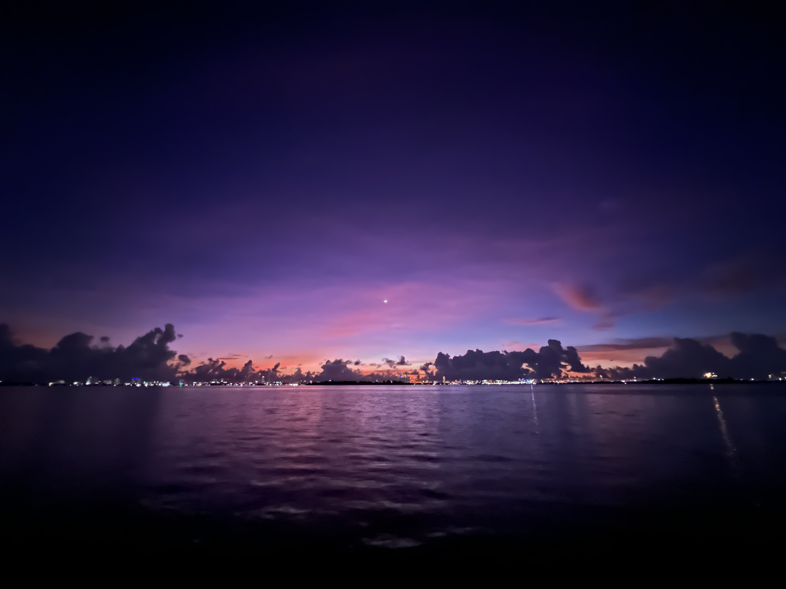 A sunset over a body of water with a sky transitioning from pink and purple to dark blue, scattered clouds, and city lights on the horizon.