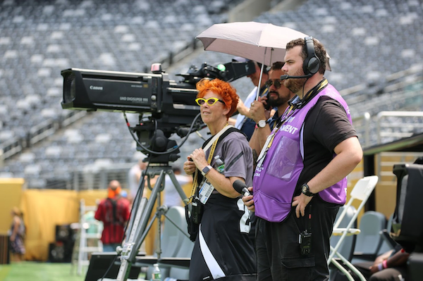 People operating a large professional video camera on a field, with one holding an umbrella for shade; they are wearing headsets and equipment, likely at a sports event.