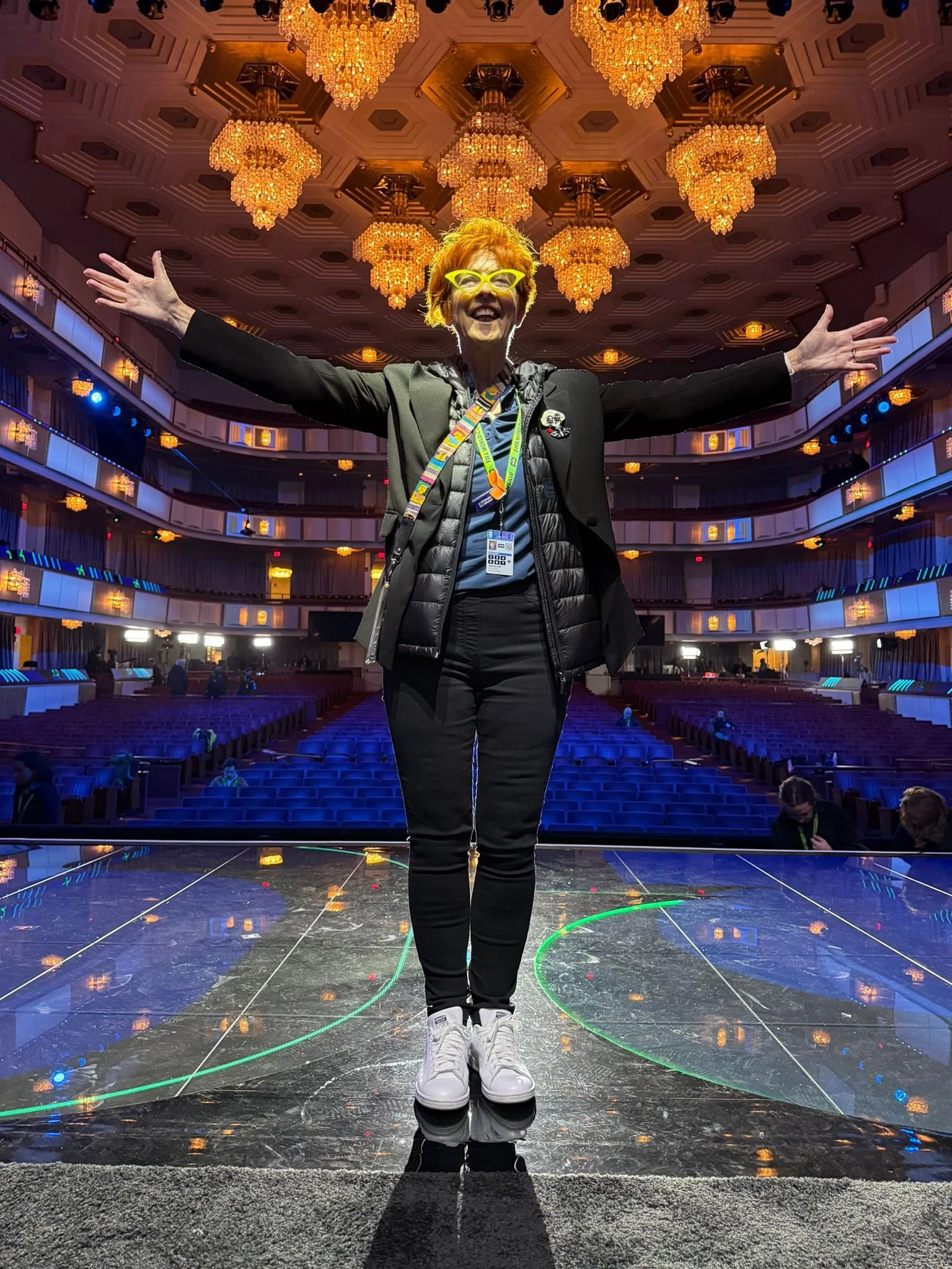 Woman standing on a stage with arms outstretched inside a theater or concert hall, with multiple ornate chandeliers hanging from the ceiling, and empty rows of seats in the background.