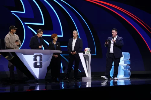 Five people standing on a TV game show stage with a futuristic blue and red backdrop, two holding microphones and one with a laptop, and a small white table with a large 3D number 3 and a spinning ball.