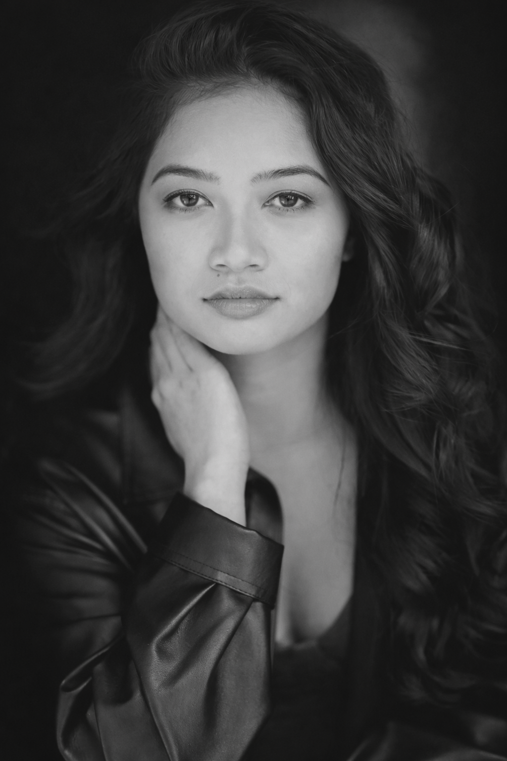 Black and white portrait of a woman with long, wavy hair, gazing into the camera with a neutral expression, resting her chin on her hand, wearing a leather jacket.