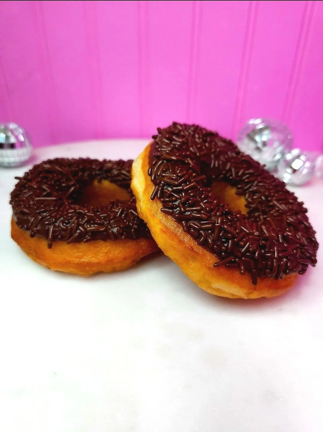 Two donuts resting against each other with chocolate frosting and chocolate sprinkles, on a white marble surface against a pink background with disco ball accents.