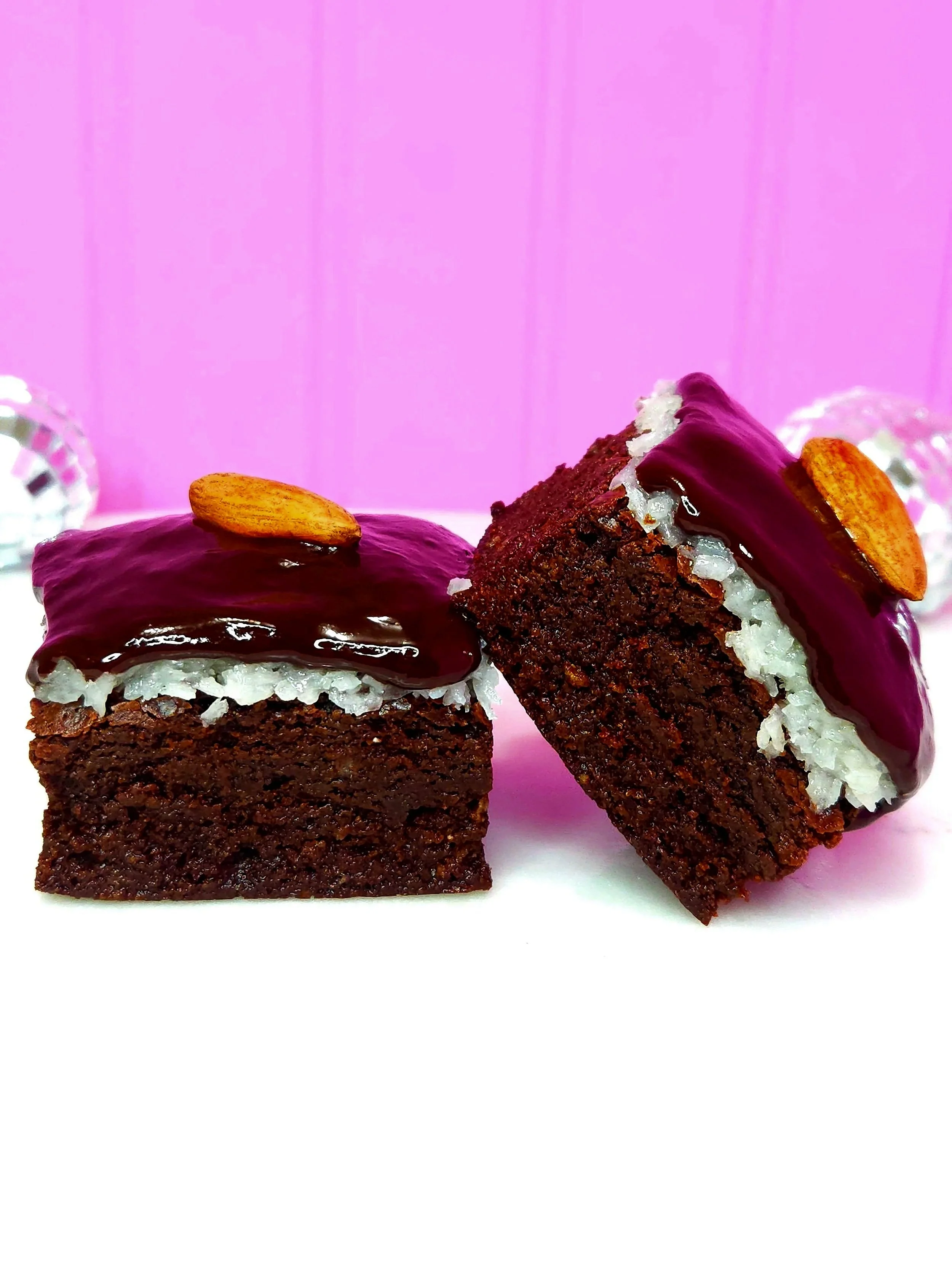 Two chocolate brownie babies with a layer of sweet coconut cream filling, and chocolate ganache icing, and an almond on top, and on a white surface with a pink background.