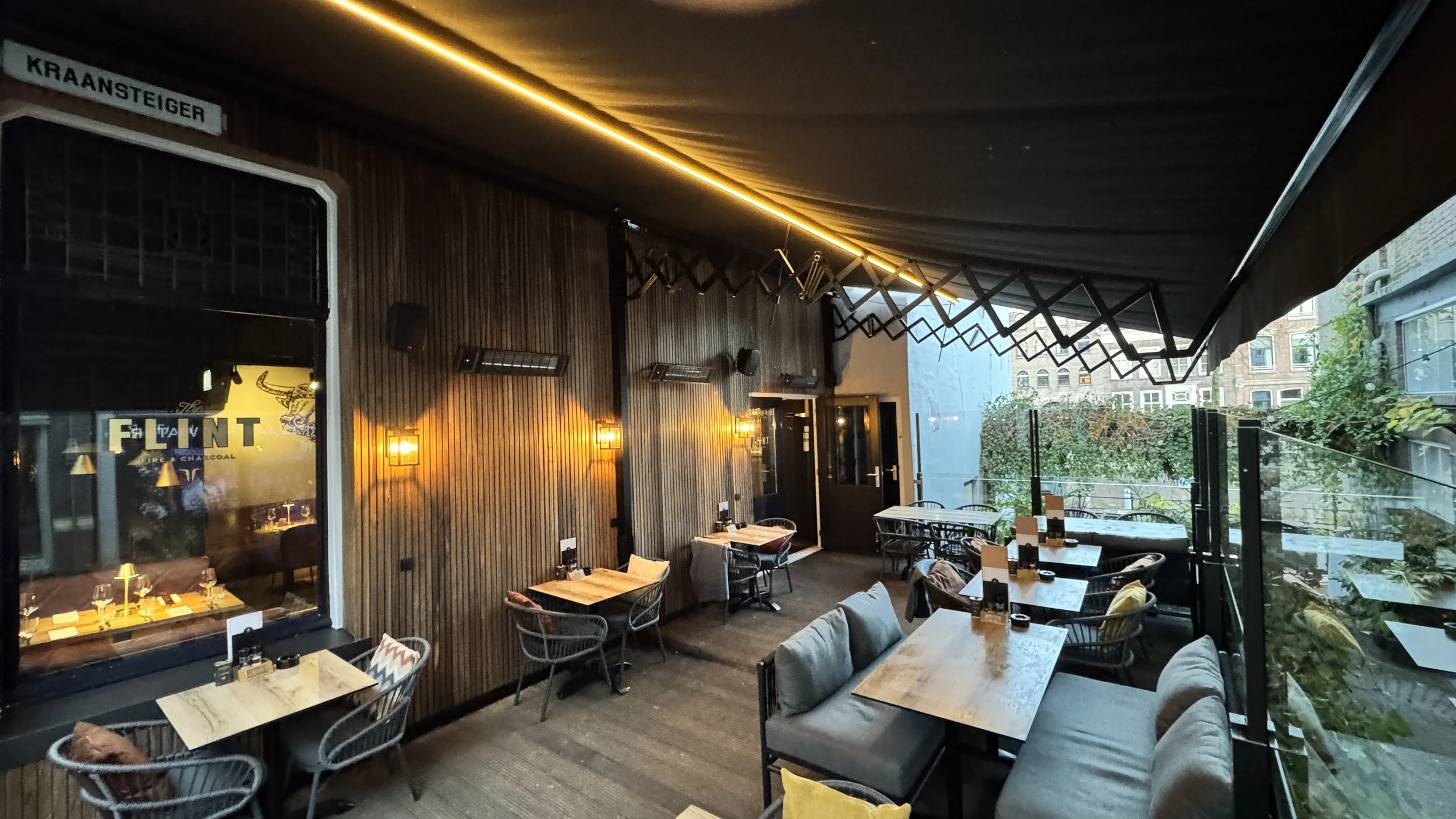 Gezellige, moderne restaurant met houten wandpanelen, warme verlichting, diverse zitplaatsen met kussens, en een glazen balkon met uitzicht op de straat en gebouwen in de achtergrond.
