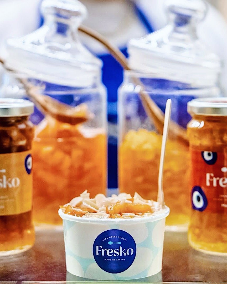 Fresko Yoghurt Bar, Athens & London. A cup of Greek yogurt with peach slices and shredded coconut in front of jars of honey with honey dippers.