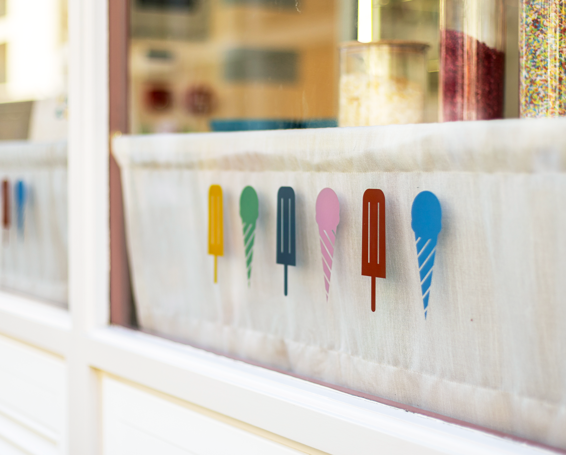 Sticks & Cones, London. Coloyrful ice lollies decoration on a white fabric banner displayed inside a window.