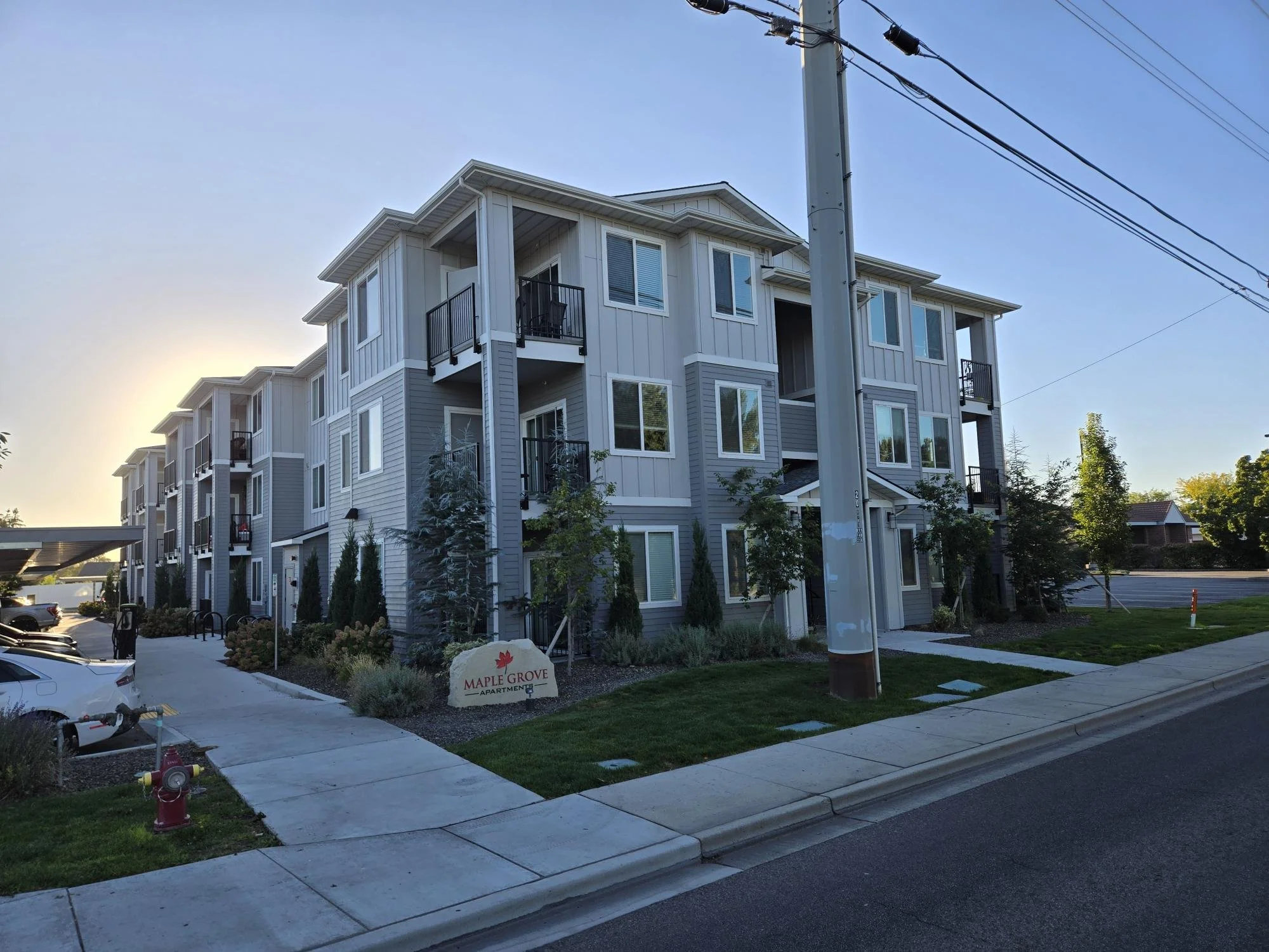 A multi-story apartment building with gray exterior and white trim, surrounded by landscaped greenery and a sidewalk. A sign reads 'Maple Grove Apartments' at the front, with cars parked nearby and power lines overhead.