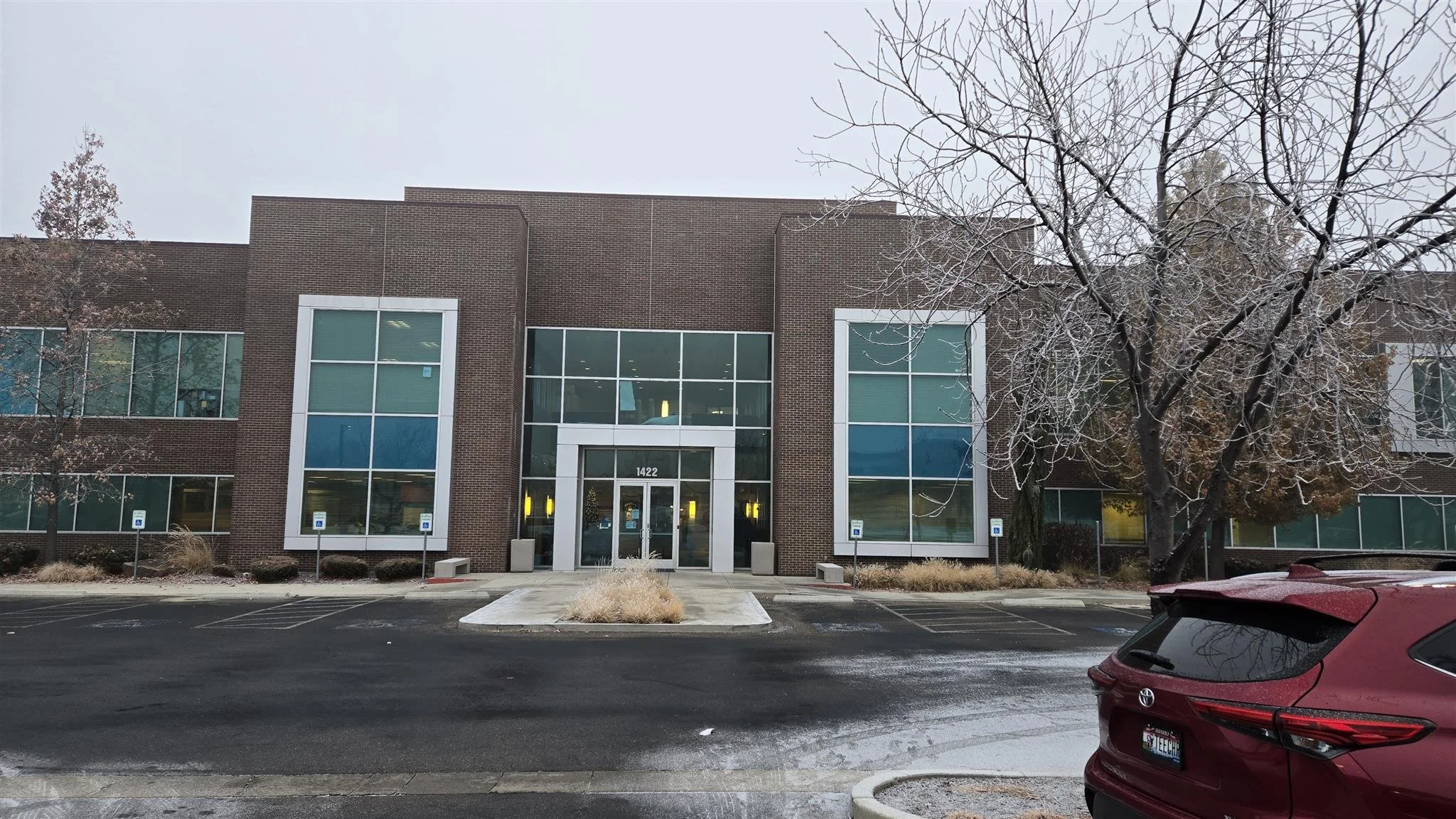 A two-story office building with large glass windows and a brick facade, numbered 1422, with a small parking lot in front and a few cars parked, including a maroon vehicle. There are some leafless trees and cloudy skies overhead.