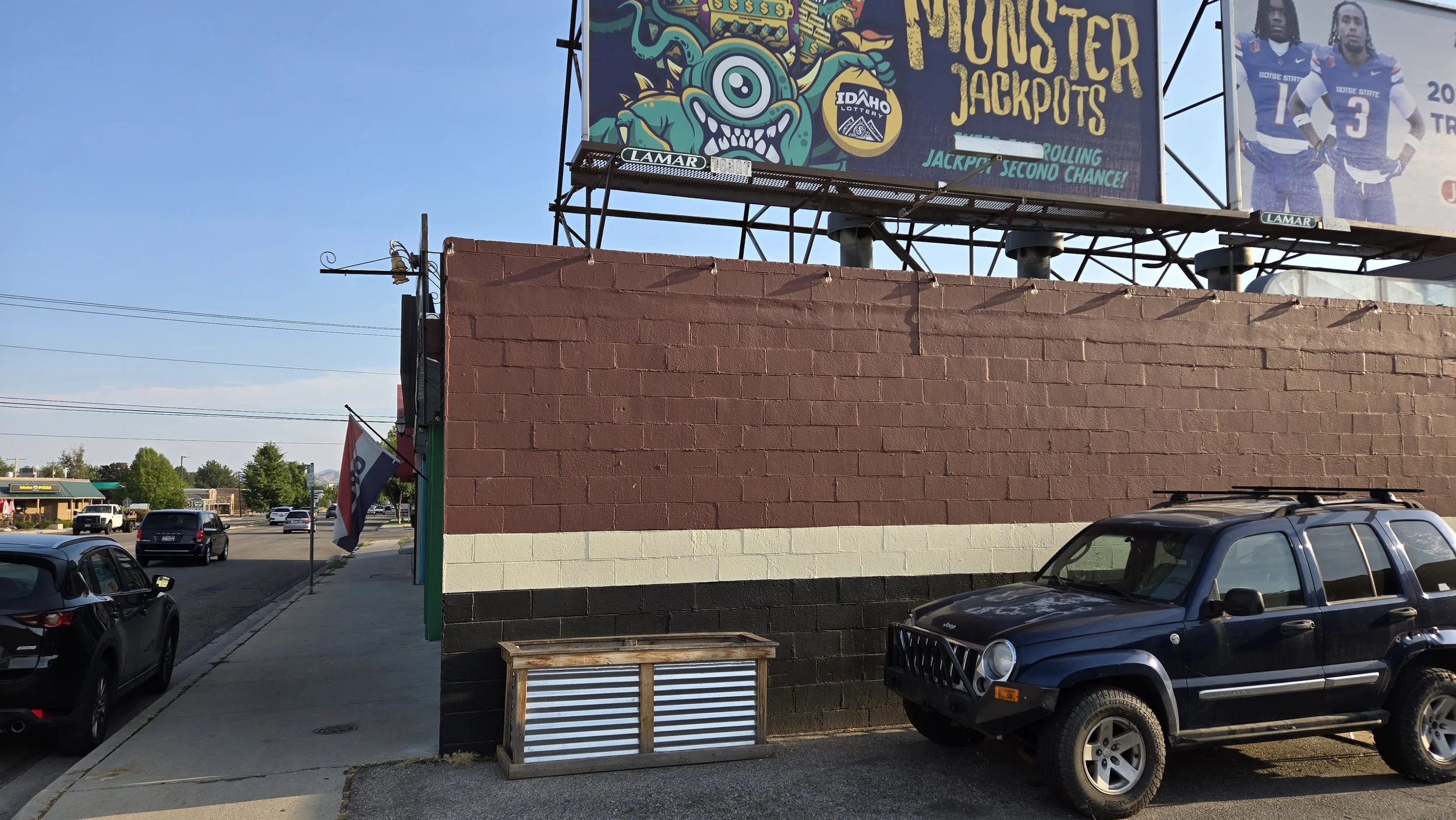 Street facing view with a brick wall, a parked black Jeep, bicycles, and a billboard advertising Idaho Lottery and Monster Jackpots, with a clear blue sky.