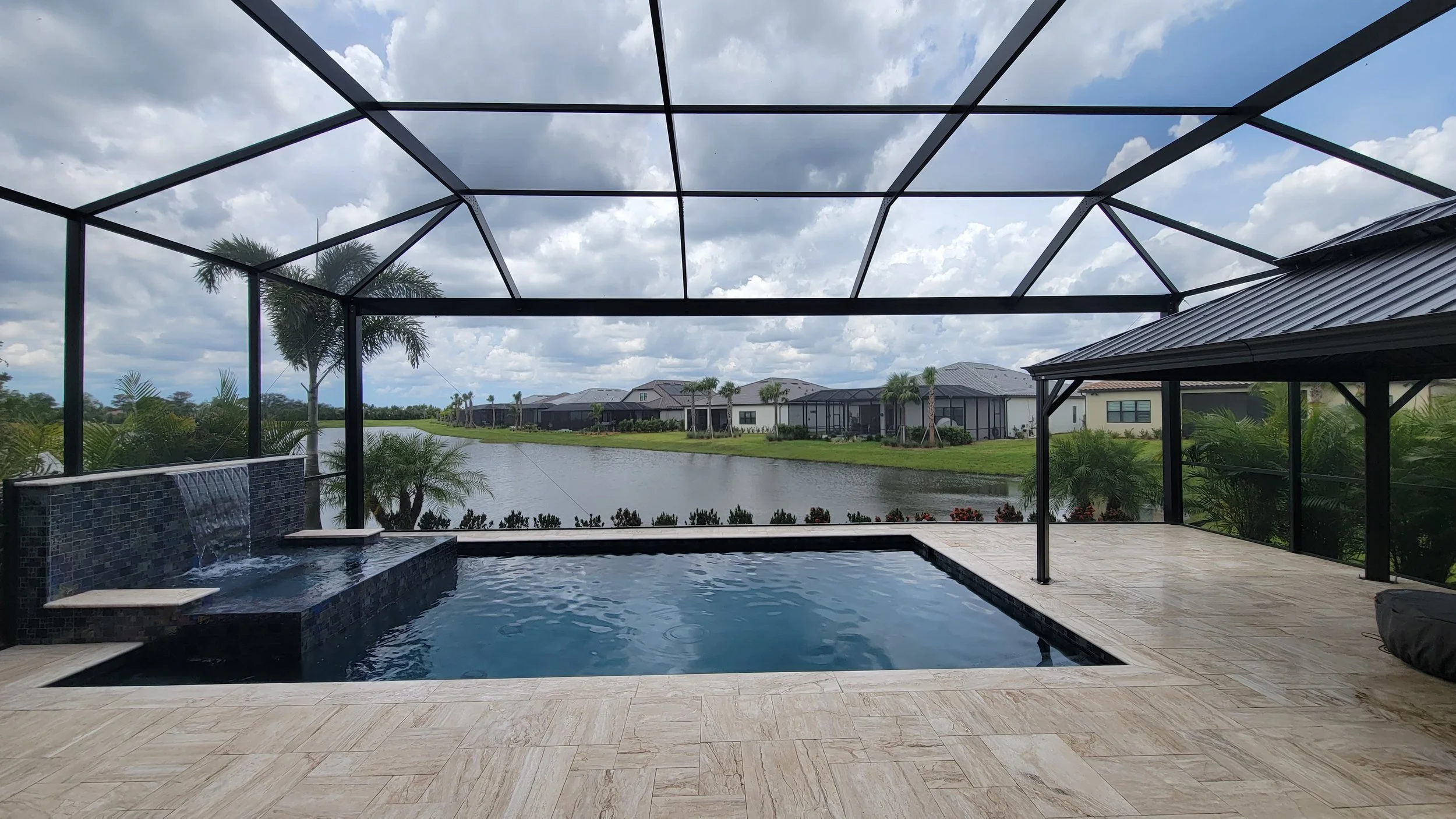 A screened-in outdoor pool area overlooking a pond with houses in the background. The pool has a small waterfall feature and beige tiled deck with some palm trees and plants around.