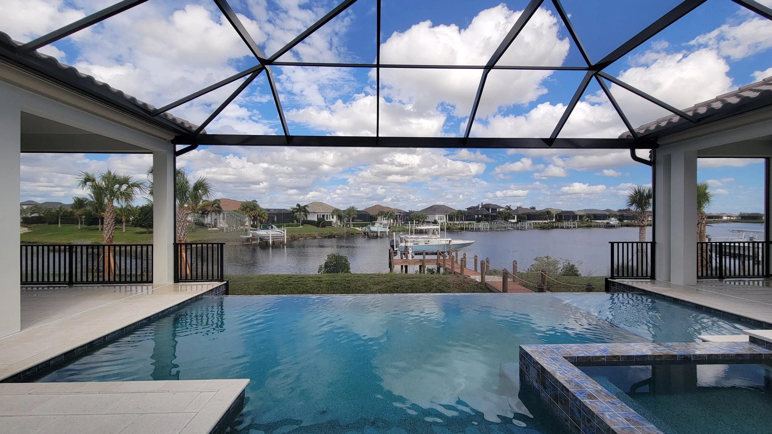 A backyard with a swimming pool overlooking a river and neighboring houses with docks, palm trees, and boats, under a partly cloudy sky.