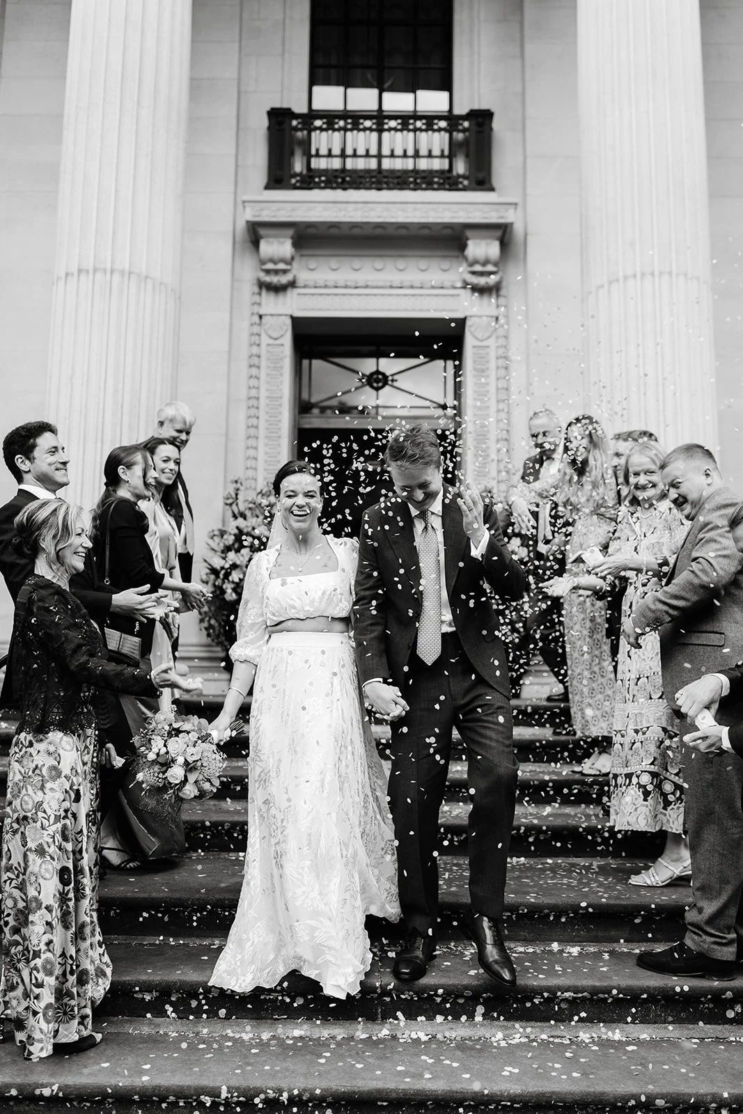 Couple celebrating outside Old Marylebone Town Hall in London, with colorful confetti falling around them, capturing a joyful wedding moment in a documentary style.