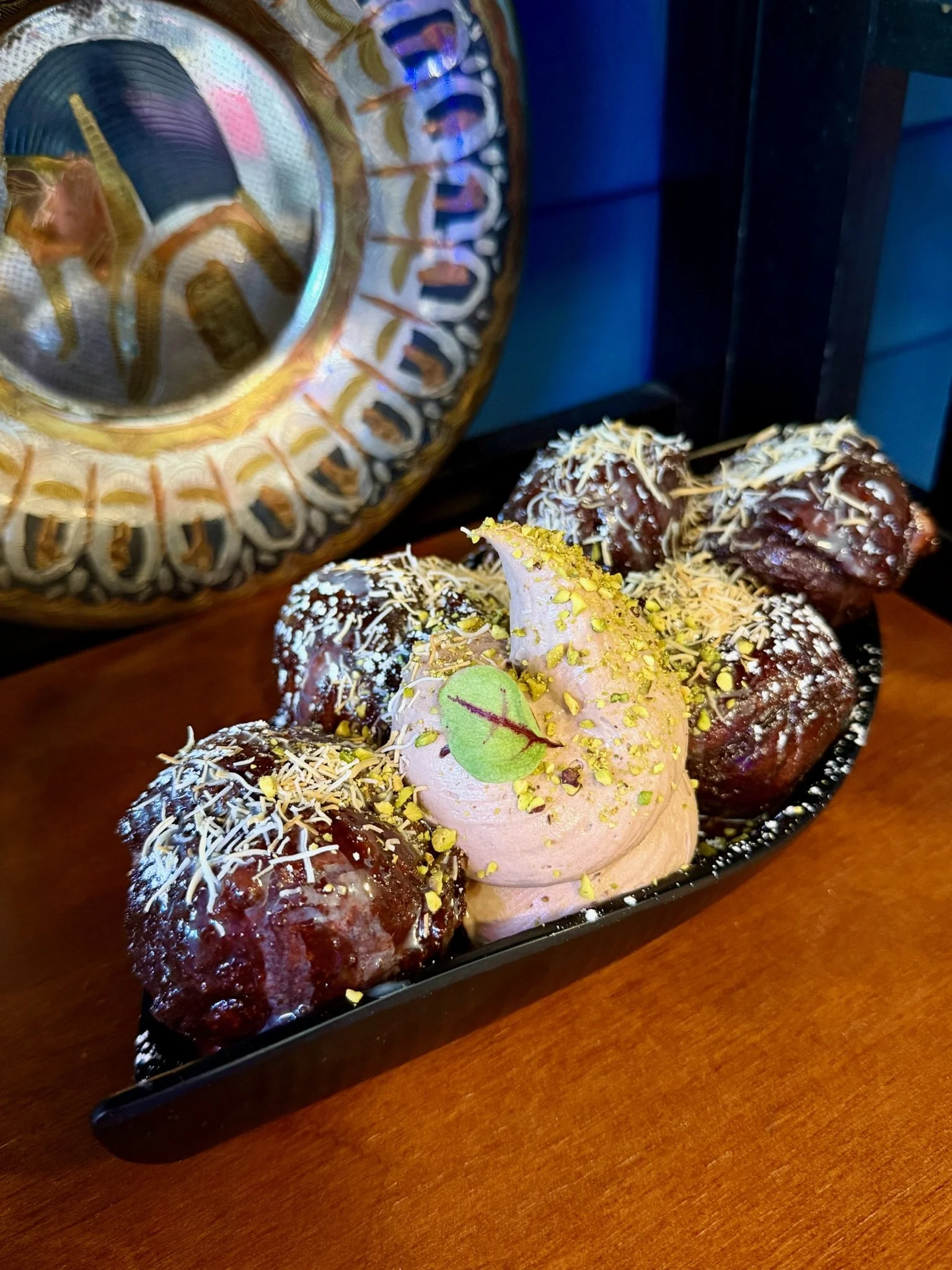 Chocolate dessert balls with shredded white chocolate, seeds, and a dollop of pink creamy topping garnished with a green leaf on a black rectangular plate.