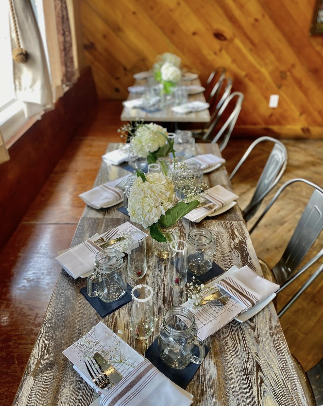 A rustic dining table set for a meal with white floral centerpieces, napkins, plates, glasses, and utensils in a cozy room with wood-paneled walls and natural light.