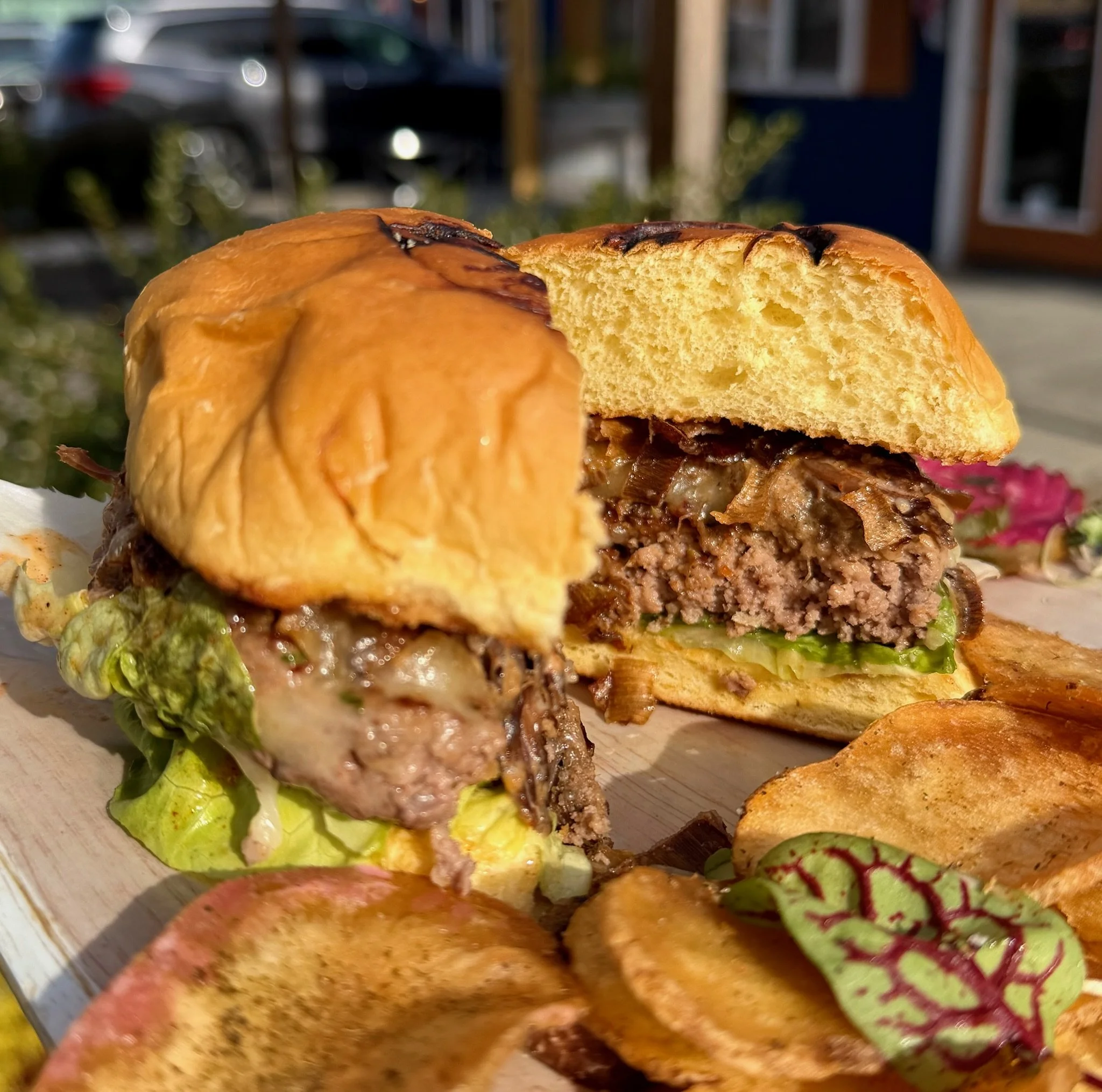 Close-up of a half-eaten burger with lettuce, cheese, beef patty, caramelized onions, and a toasted bun, with fries and pickles nearby, outdoors in sunlight.