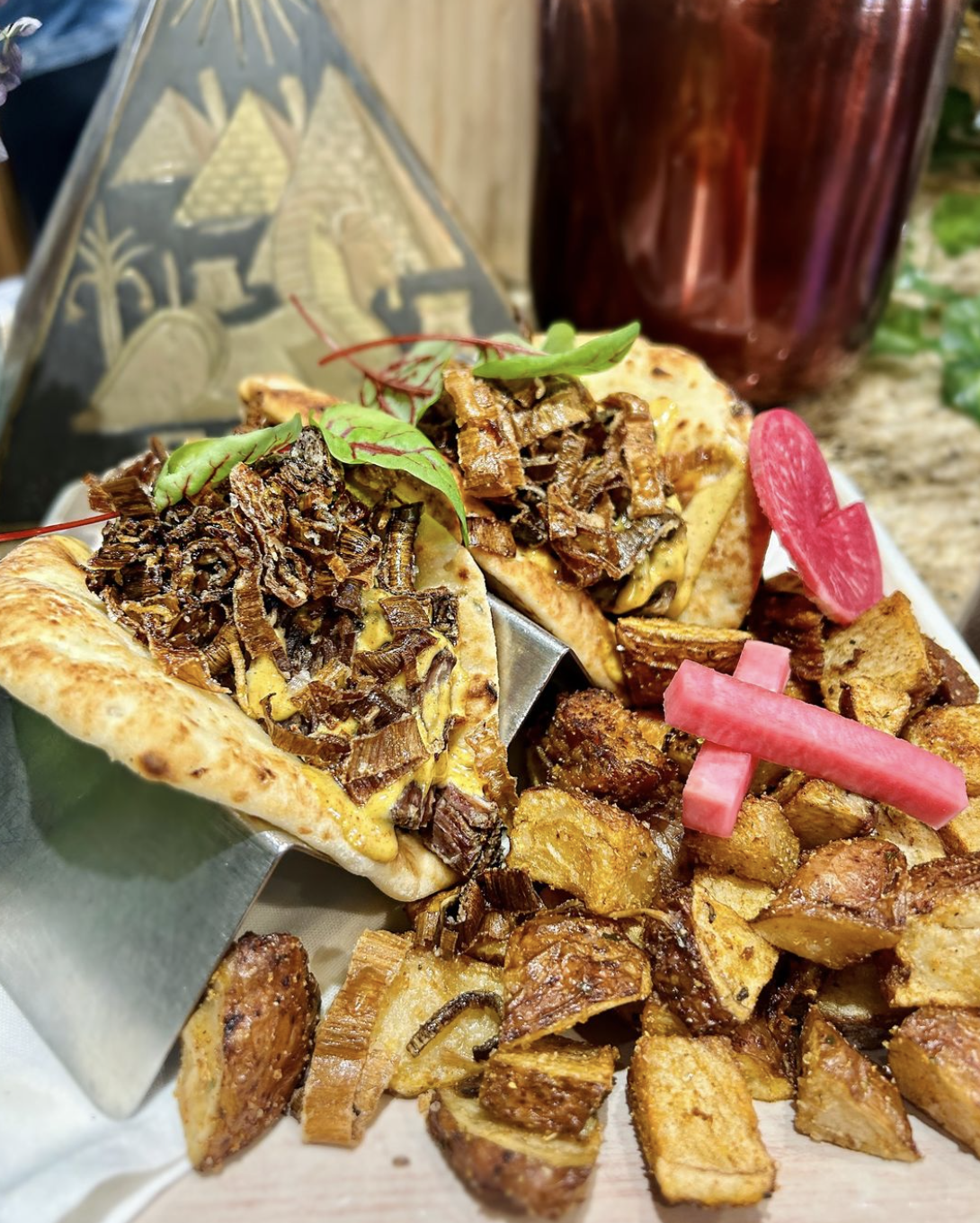 A plate with two tacos filled with shredded beef, lettuce, and cheese, accompanied by roasted sweet potatoes and pickled radishes, with a dark beverage in the background.