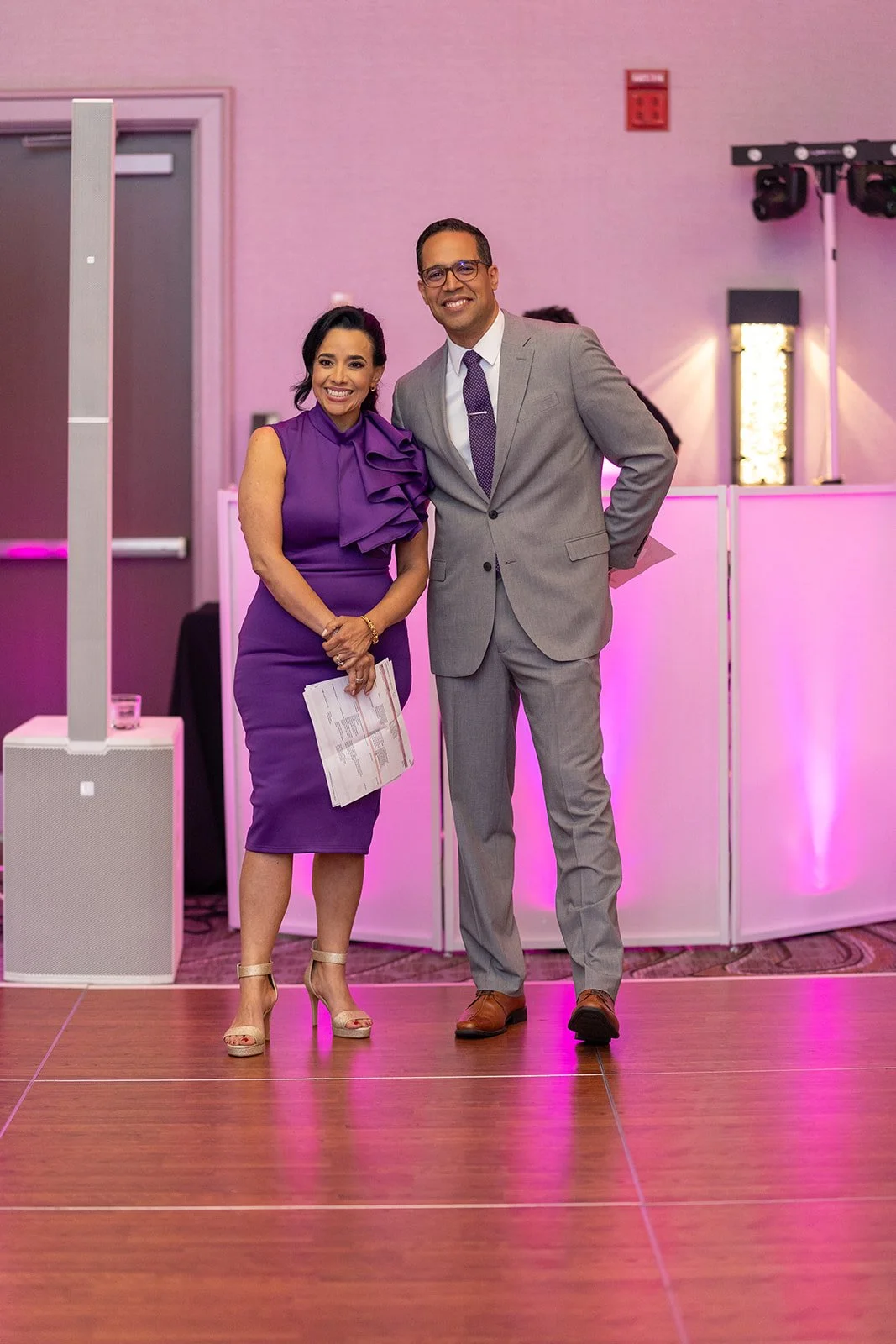A woman in a purple dress and a man in a gray suit standing together at an event, smiling, with pink lighting and event equipment in the background.
