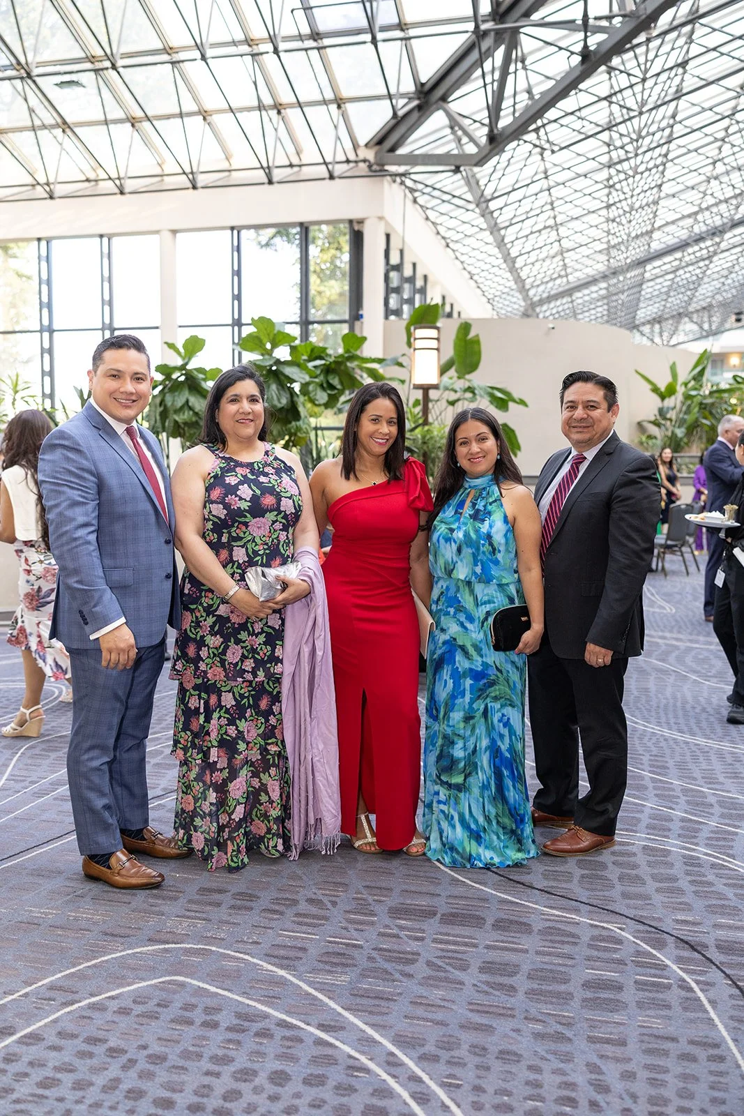 Group of six people posing at an indoor event, dressed in formal attire, with a modern glass ceiling and large green plants in the background.